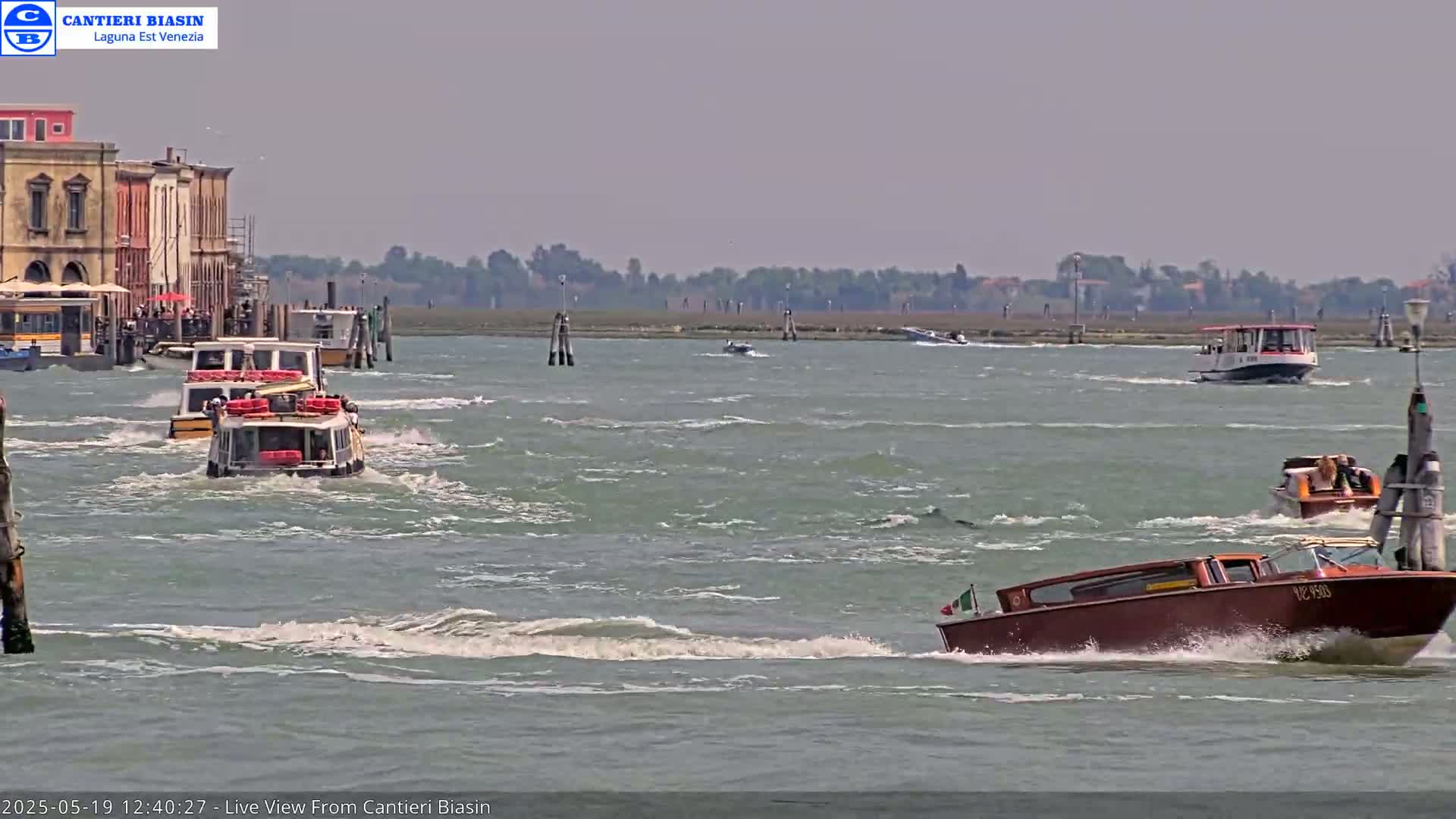 Several water taxis and smaller motorboats navigate a waterway near a city waterfront on a slightly hazy day.