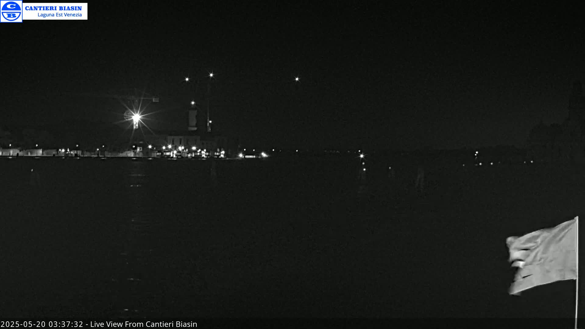 A nighttime view of a waterfront area with a few lights visible in the distance, a flag waving in the foreground, under a clear, dark sky.