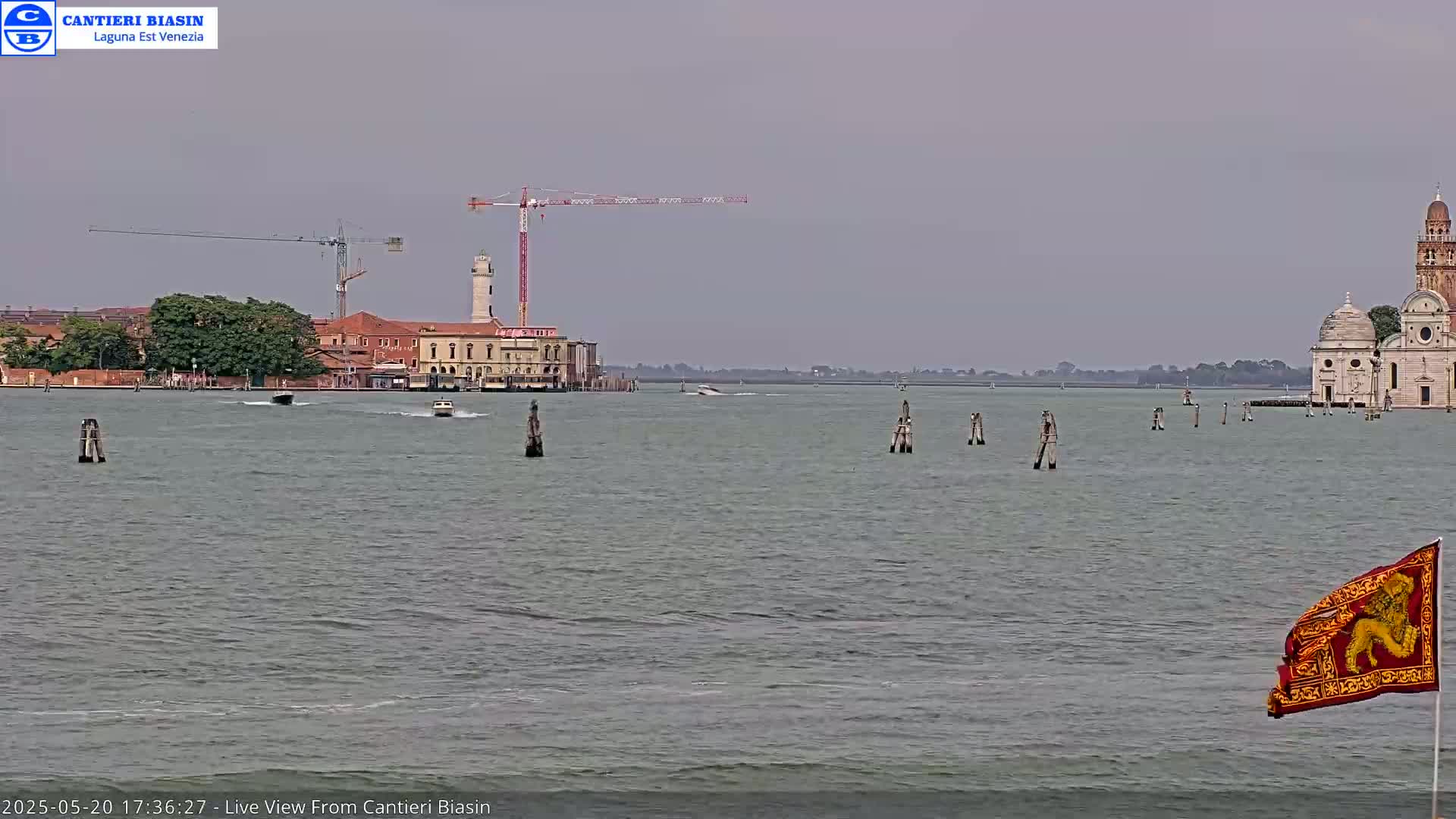 A calm, overcast day shows a Venetian waterway with several boats, a crane on a building, and a church in the distance.