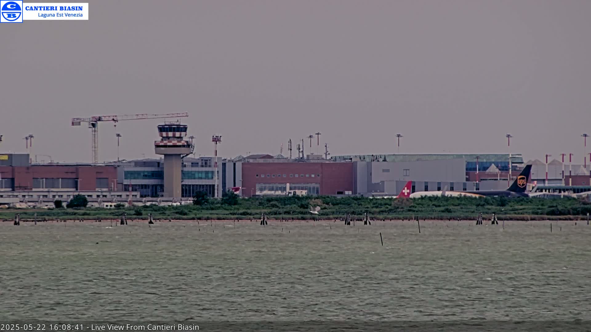 A long shot of an airport with a control tower and airplanes, viewed across a body of water under an overcast sky.