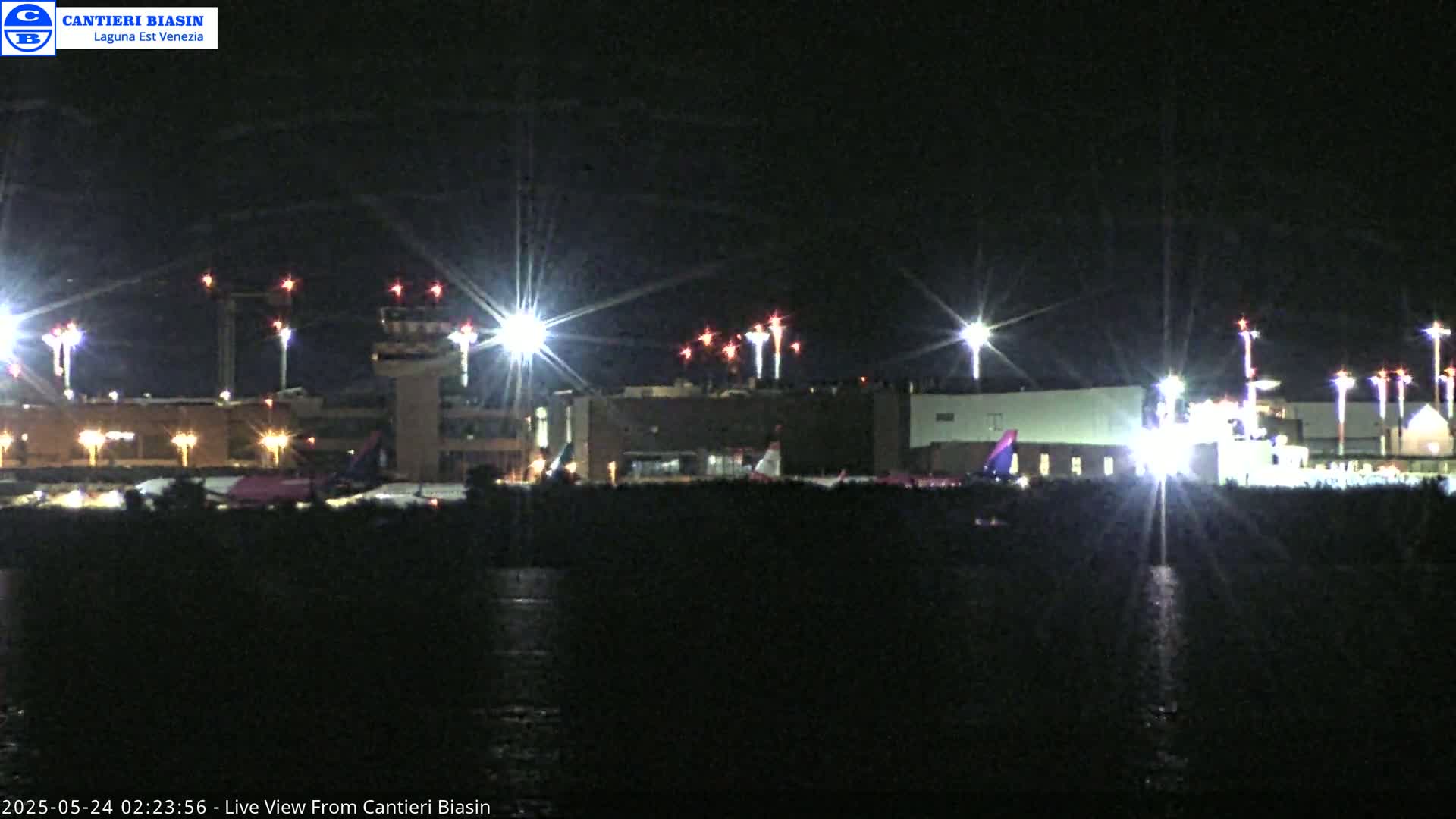 A brightly lit airport at night is seen from across a body of dark water.