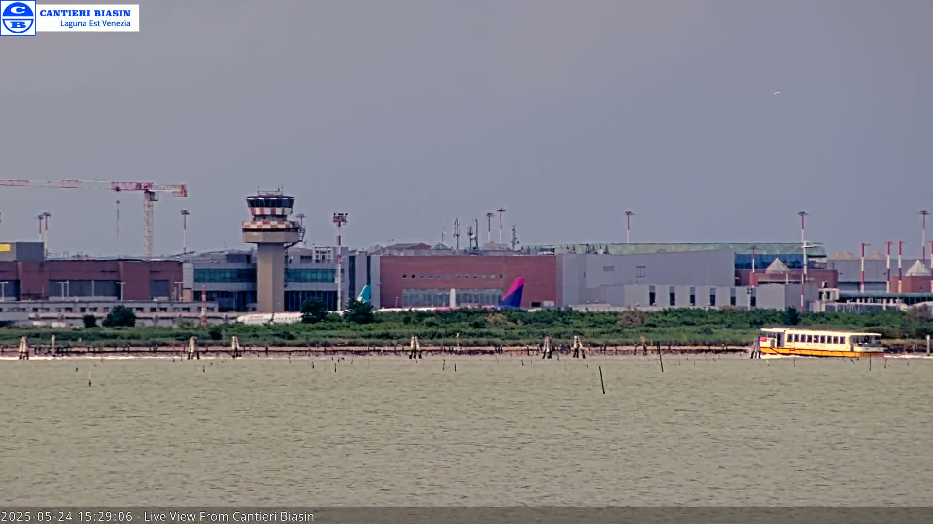 A distant view of an airport with a control tower and several buildings, partially obscured by hazy weather conditions, is seen across a body of water where a yellow ferry is present.