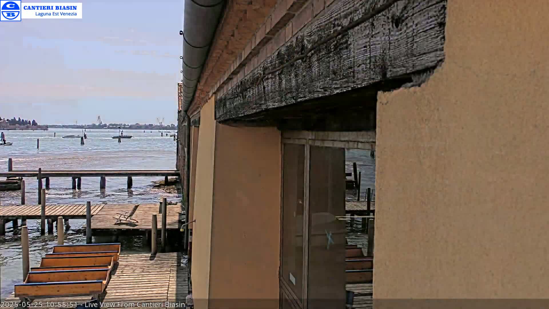 A partly sunny day reveals a waterfront scene with several boats on calm water, wooden docks, and a tan building with a large dark wood beam.