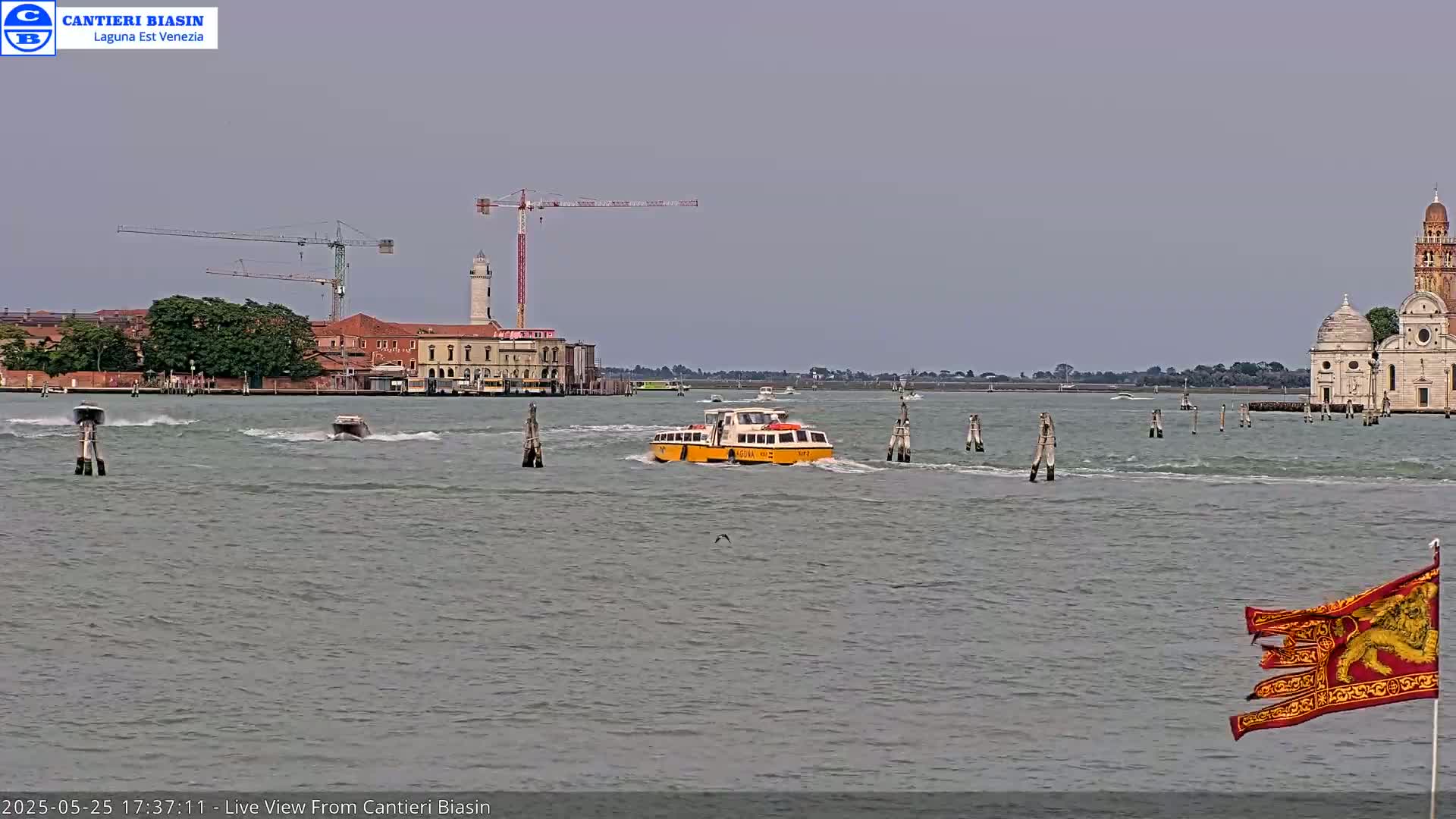 A yellow water taxi travels across a gray-green body of water under an overcast sky, past a distant island with buildings and a church, and construction cranes.
