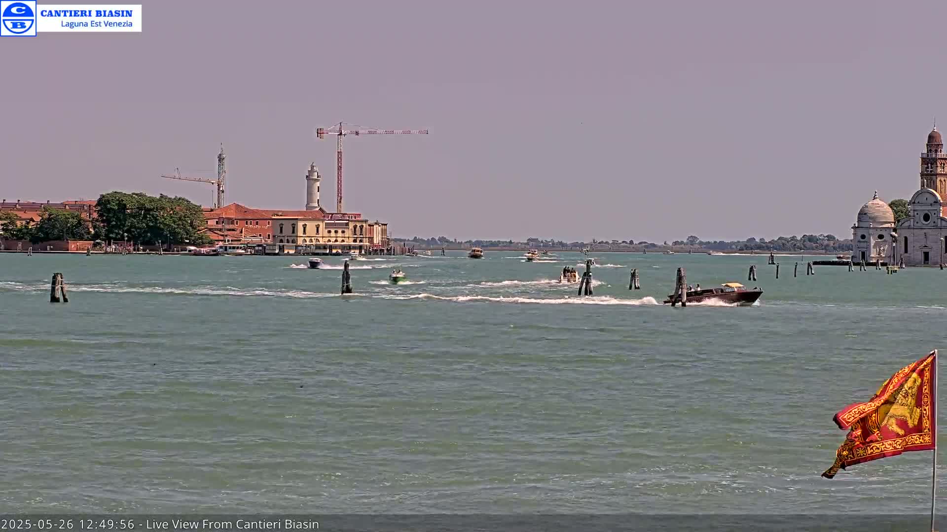 Several motorboats travel across a calm body of water on a sunny day, past buildings and a church in the distance, with a construction crane visible on the left.