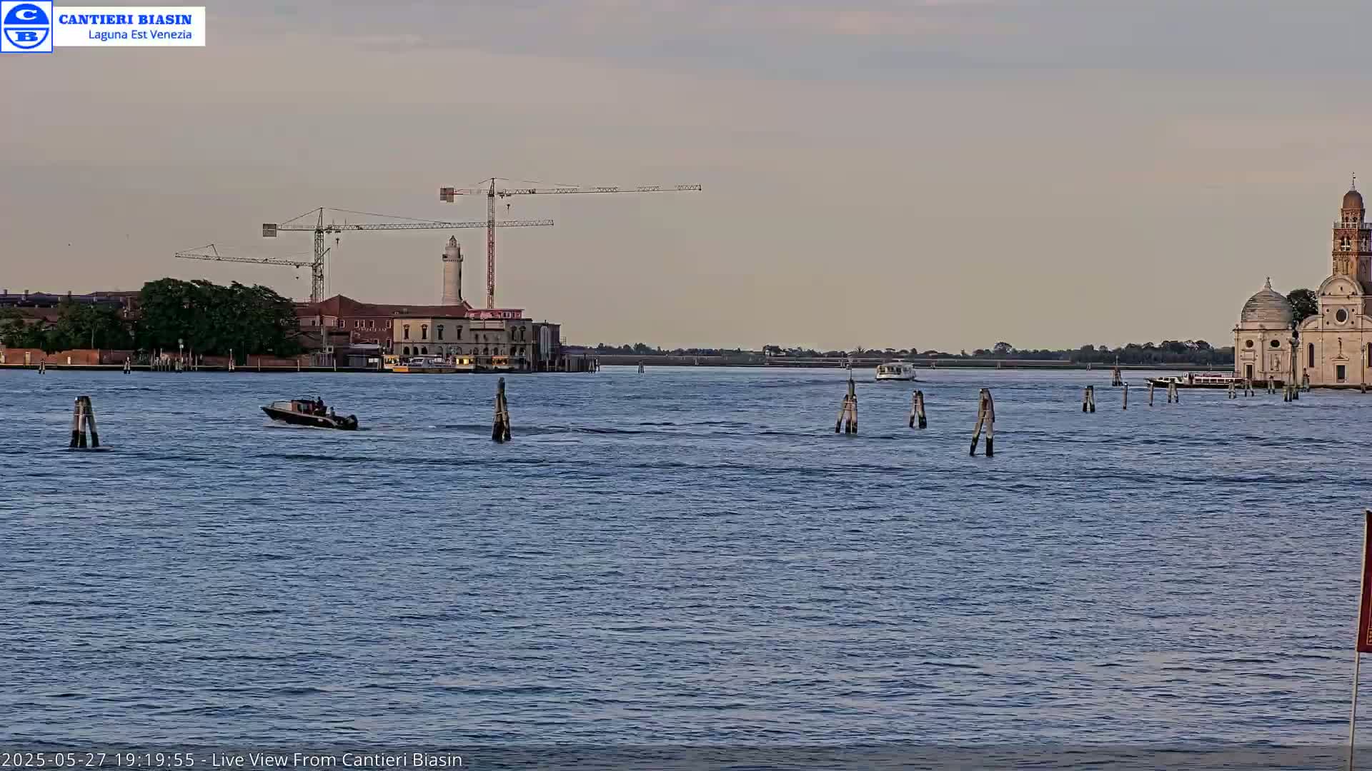 A calm body of water shows a small motorboat, two construction cranes, a lighthouse, a church, and several boats in the distance under a dusky sky.