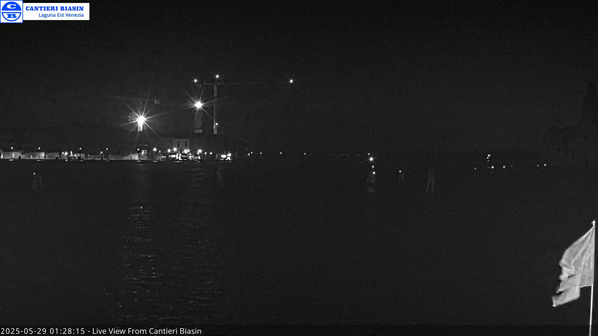 A nighttime, low-light view of a waterfront area with several lights illuminating buildings and construction cranes across a body of dark water.