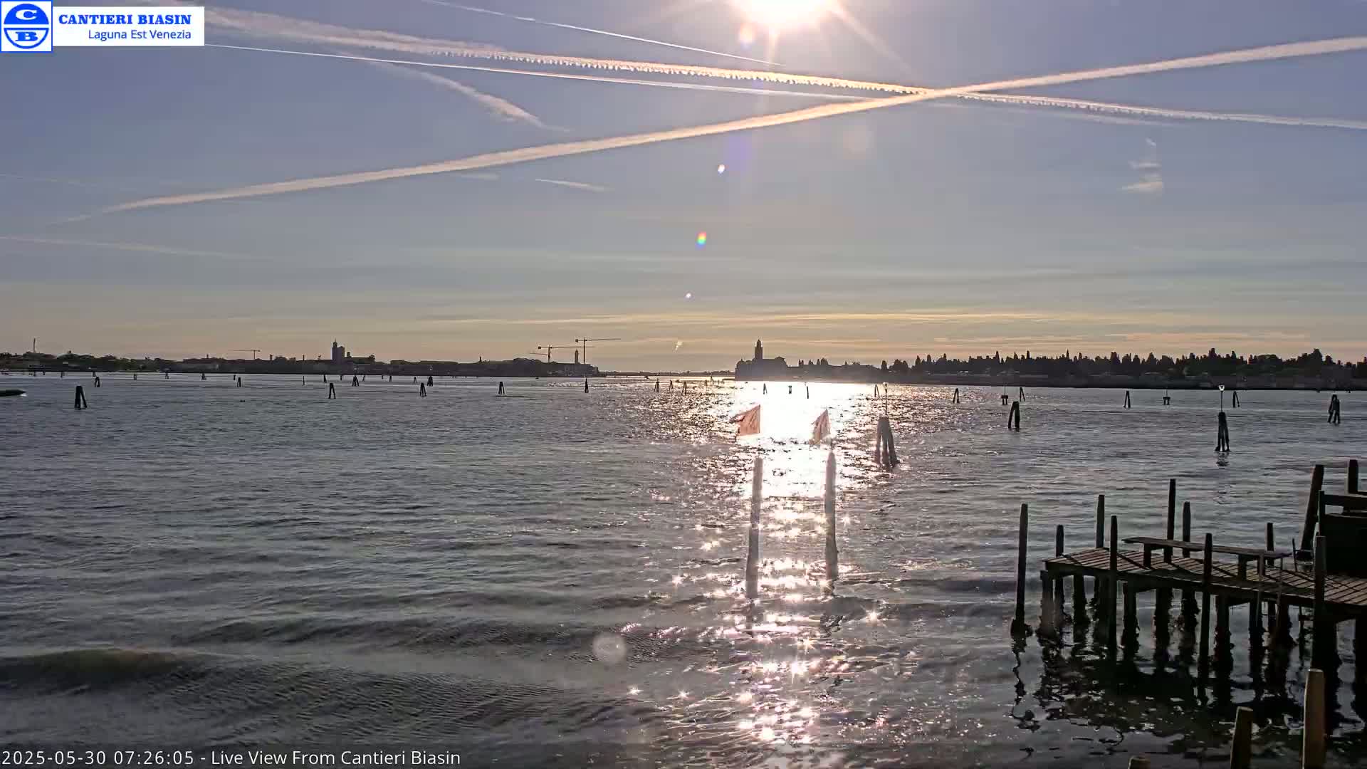 A calm body of water reflects the sun's bright light, with a distant cityscape visible under a mostly clear sky marked by contrails.