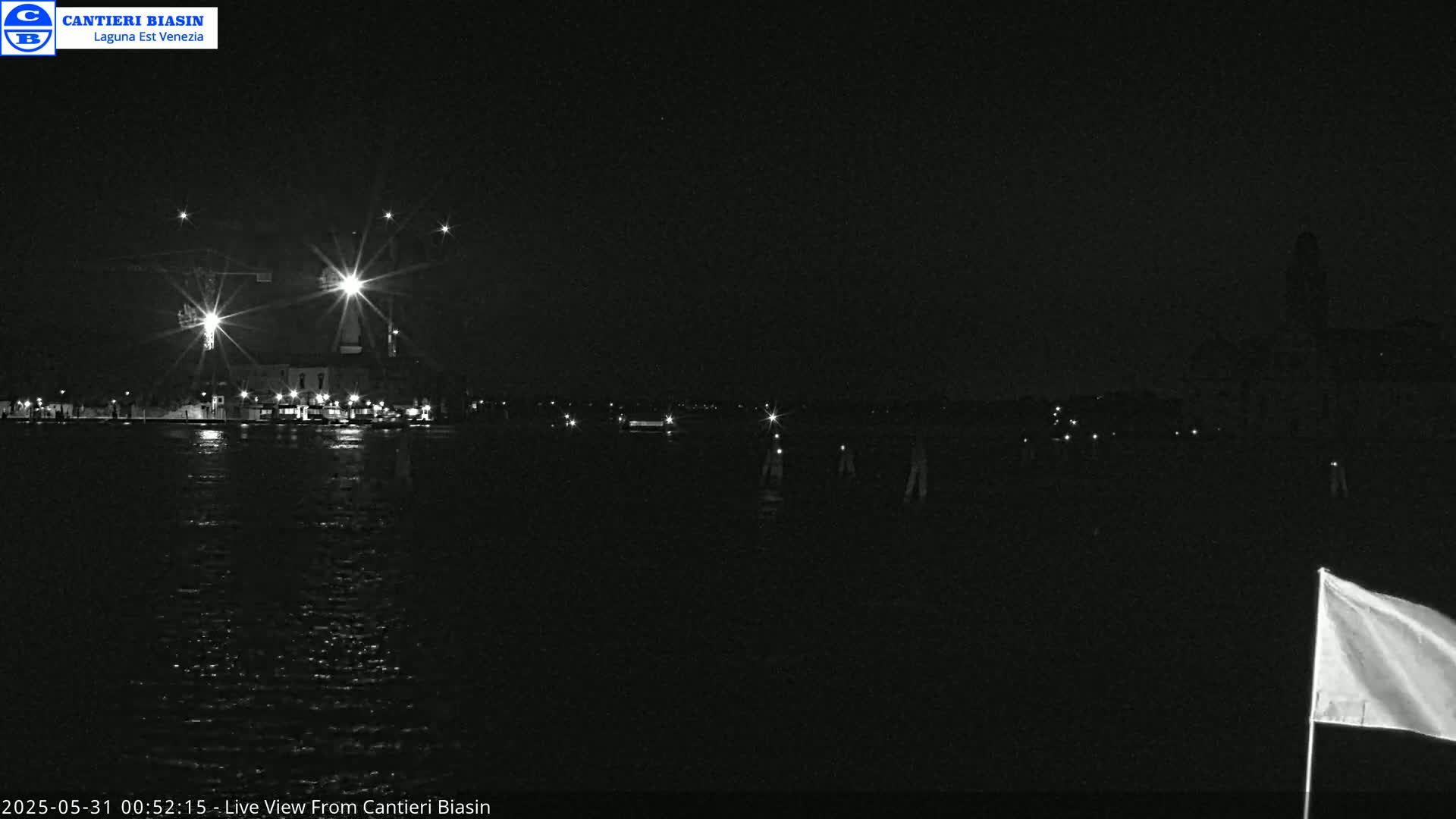 A nighttime view of a dark body of water with dimly lit buildings and a few scattered lights across the far shore, a white flag visible in the lower right corner.