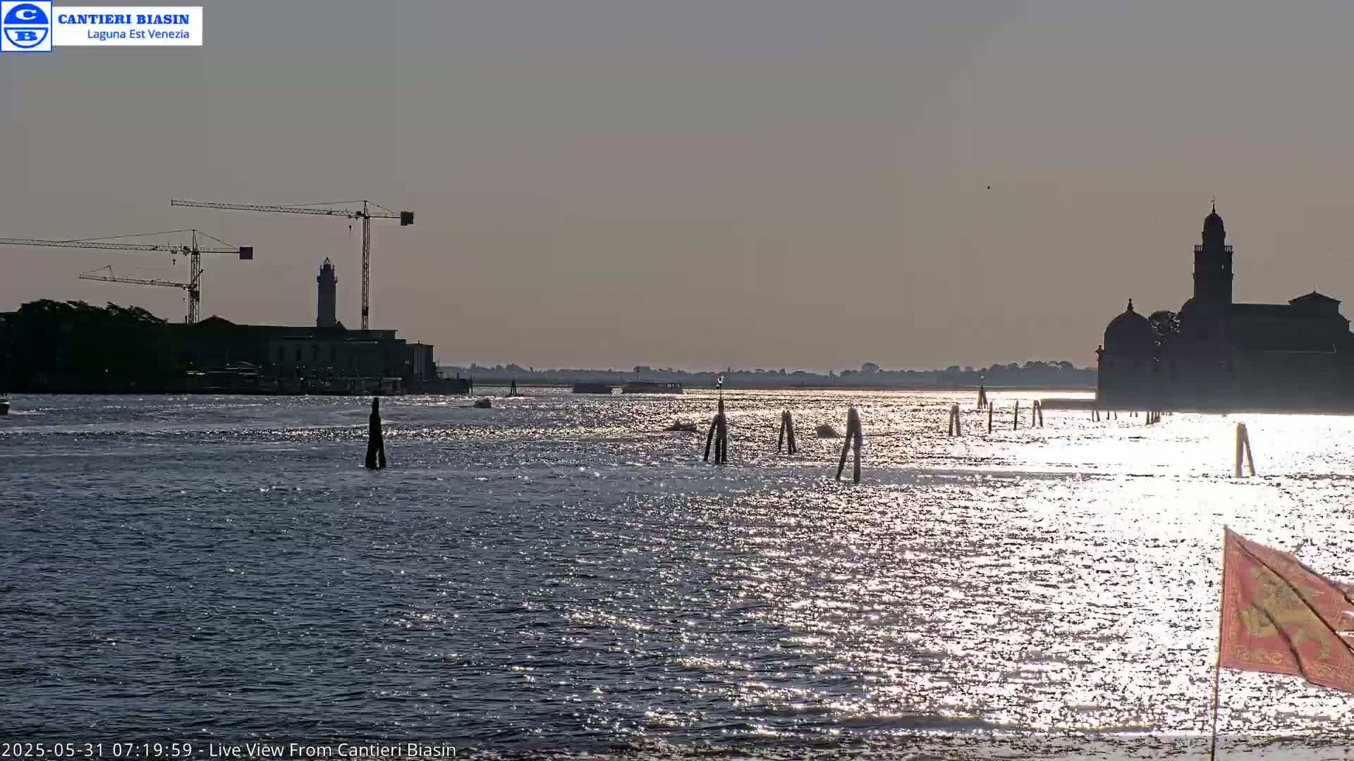 A calm body of water reflects sunlight, with silhouetted buildings and construction cranes visible in the distance under a clear sky.