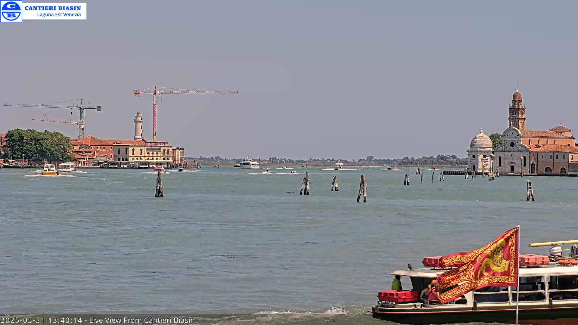 A waterway scene on a sunny day shows several boats, construction cranes, a lighthouse, and a church complex in the distance.