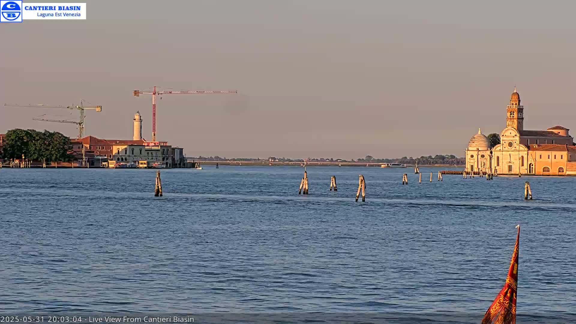 A calm body of water shows a distant island with a church and buildings, construction cranes on the left, and several wooden posts in the water.