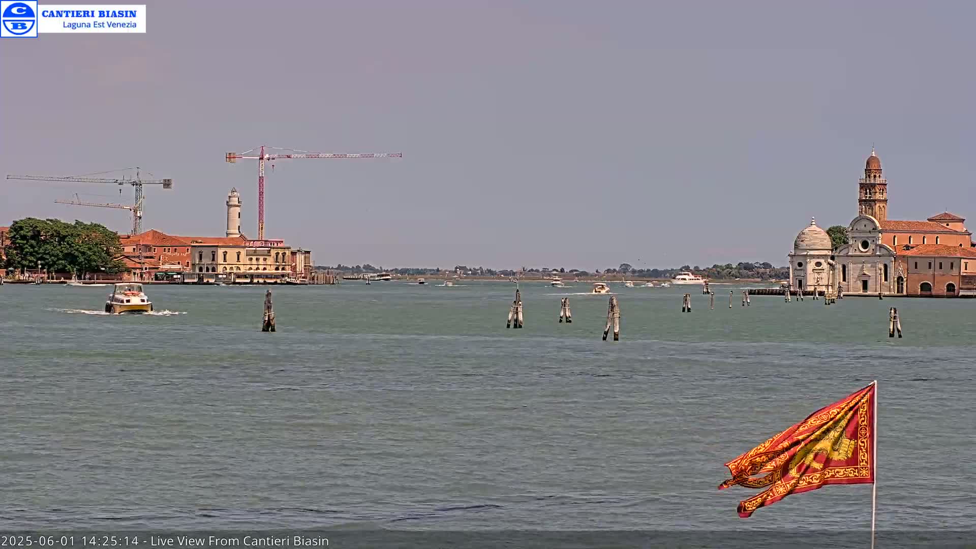 A calm body of water shows several boats, construction cranes, buildings on islands, and a red flag with a golden design blowing in the breeze under a hazy sky.