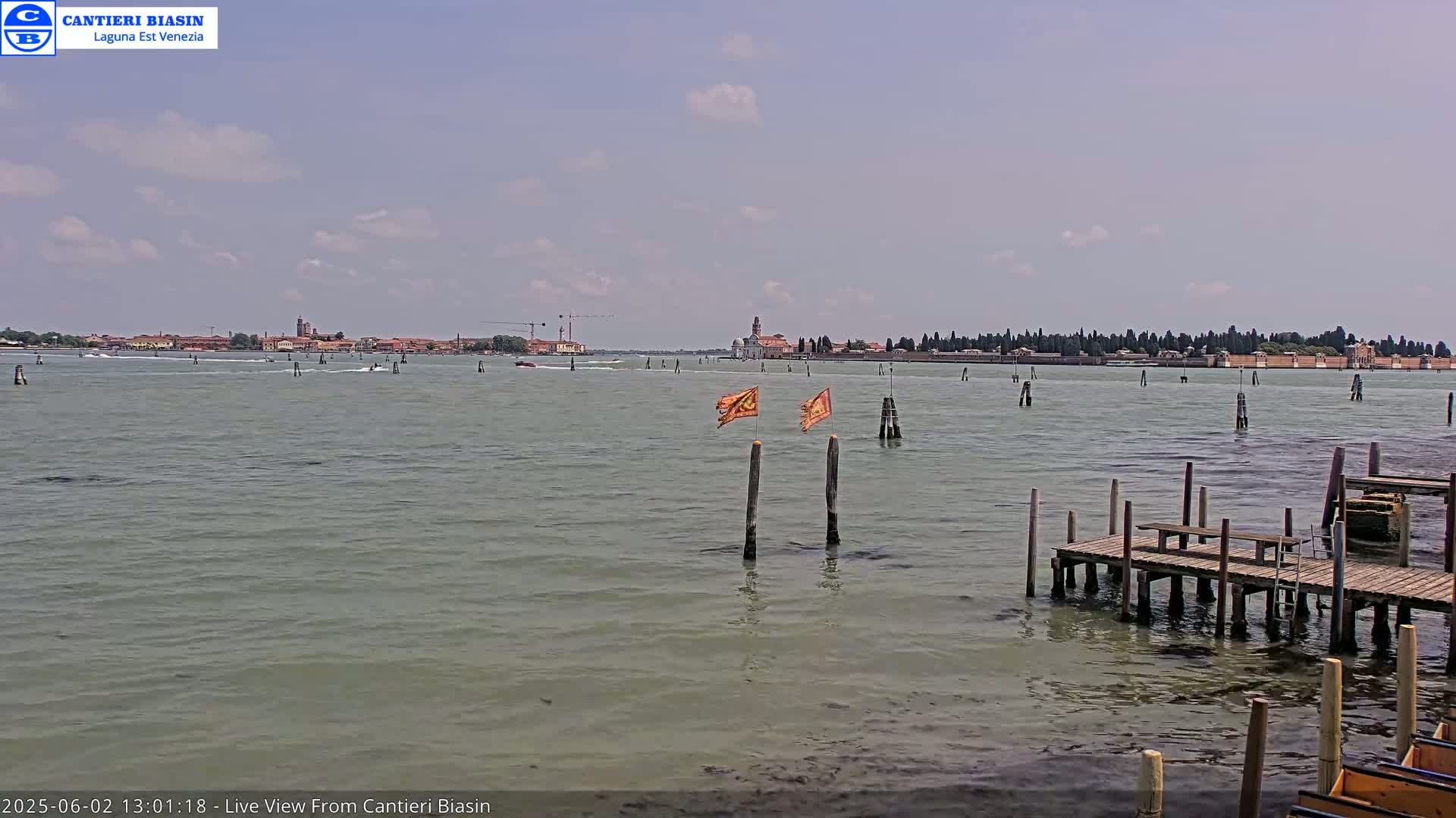 A calm, partly cloudy day reveals a body of water with several boats, wooden pilings, and a distant island with buildings visible on the horizon.
