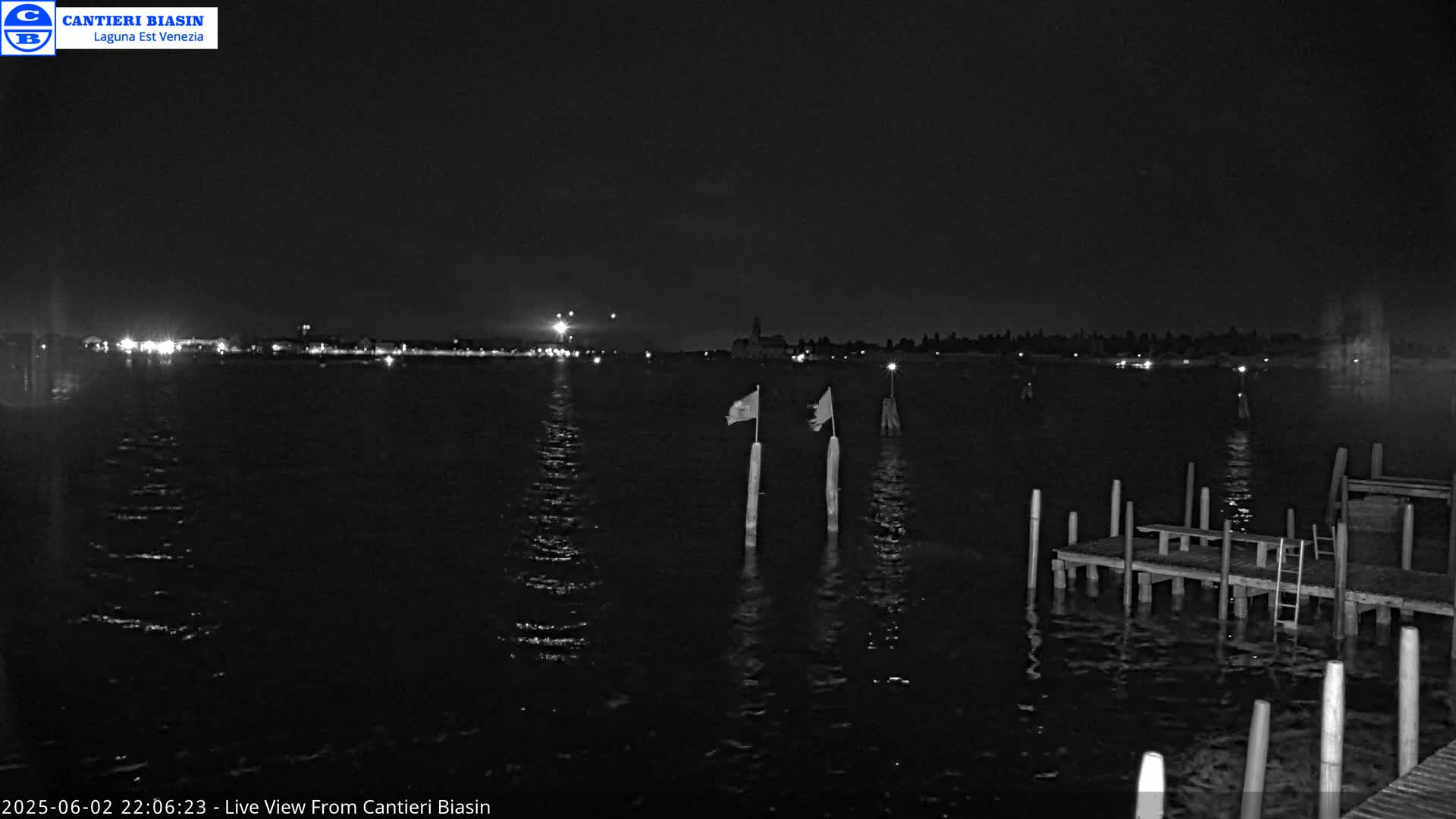 A nighttime view of a calm body of water with a distant shoreline showing lights, two flagged posts in the foreground, and a wooden dock to the lower right.