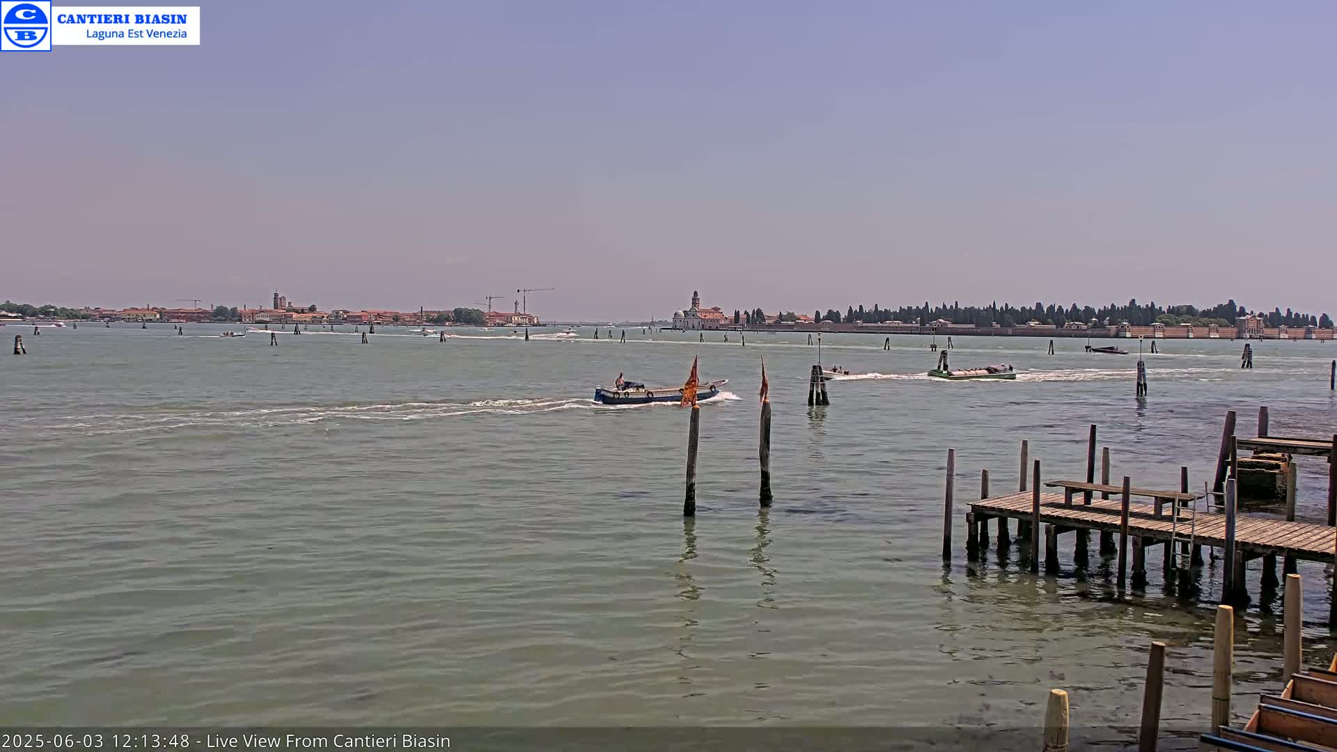 A calm body of water with several small boats, a distant island with buildings, and a wooden dock in the foreground under a clear sky.