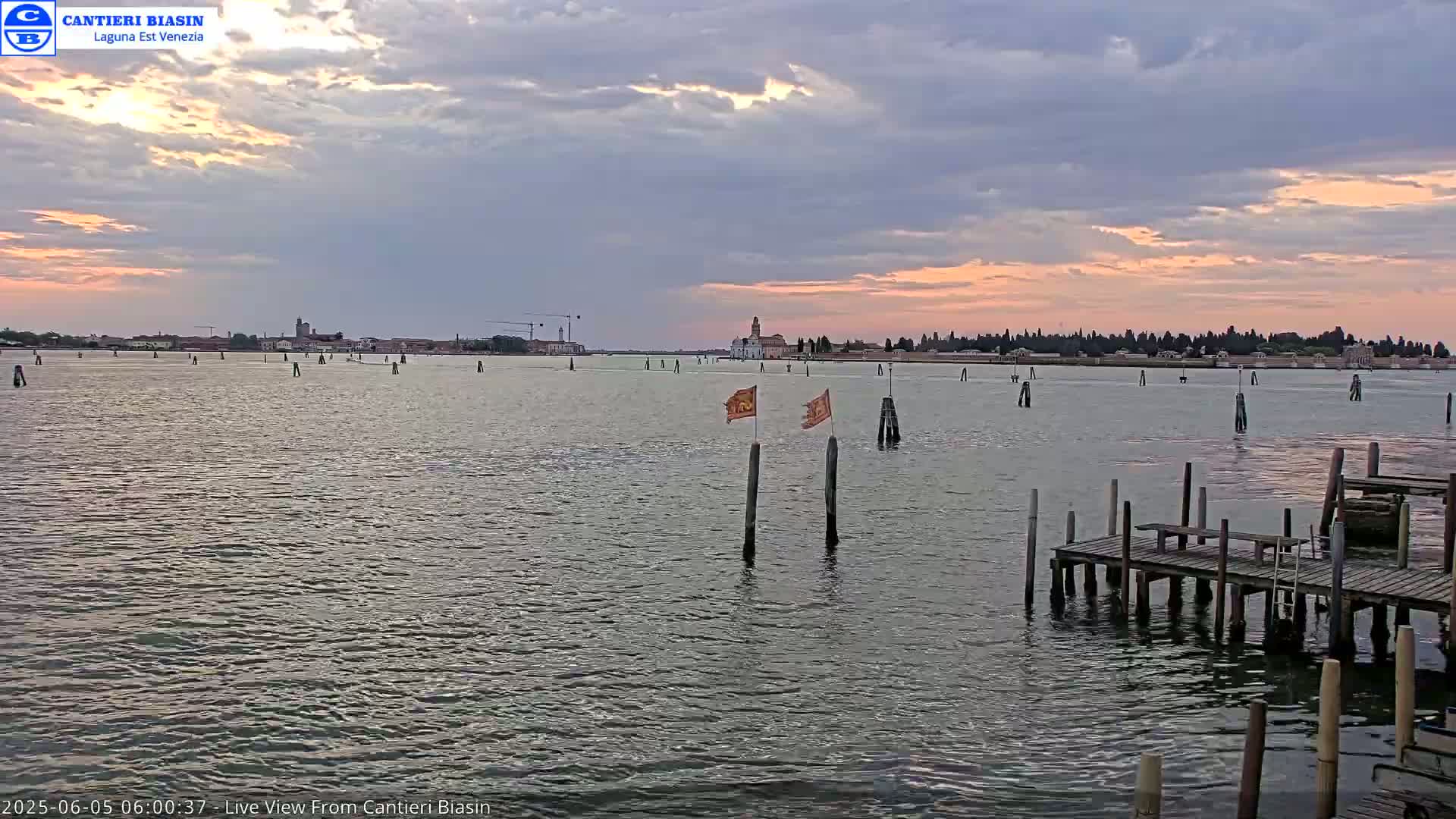 A calm body of water with a distant cityscape under a partly cloudy sky showing the colors of sunrise.