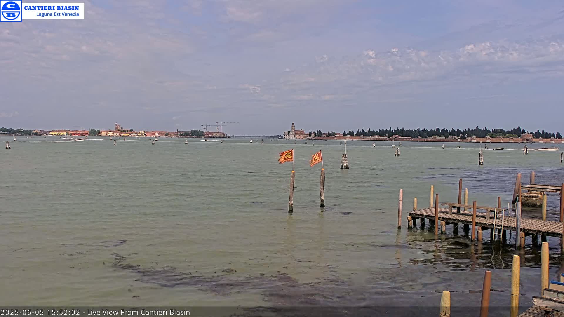 A calm, overcast day shows a view of a body of water with a distant island and a small dock in the foreground, featuring two flags on posts.
