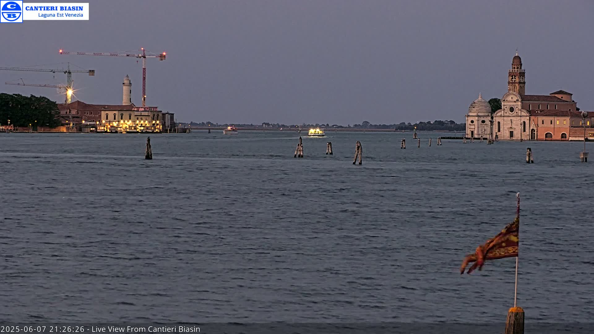 A body of water at dusk shows a building with a lighthouse and a church in the distance, with construction cranes and a boat visible.
