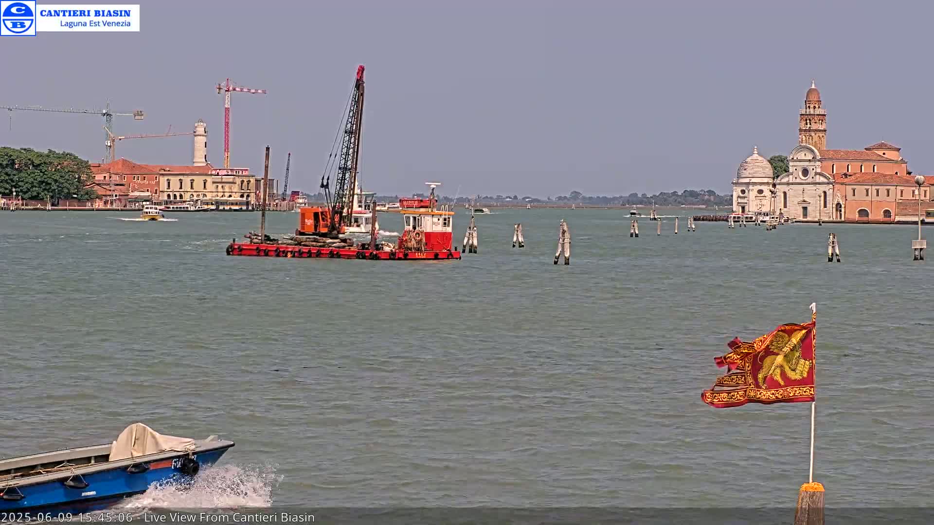 A dredge boat on calm water passes by a church and other buildings on a sunny day, with a small motorboat in the foreground and a tattered flag on a post in the lower right.