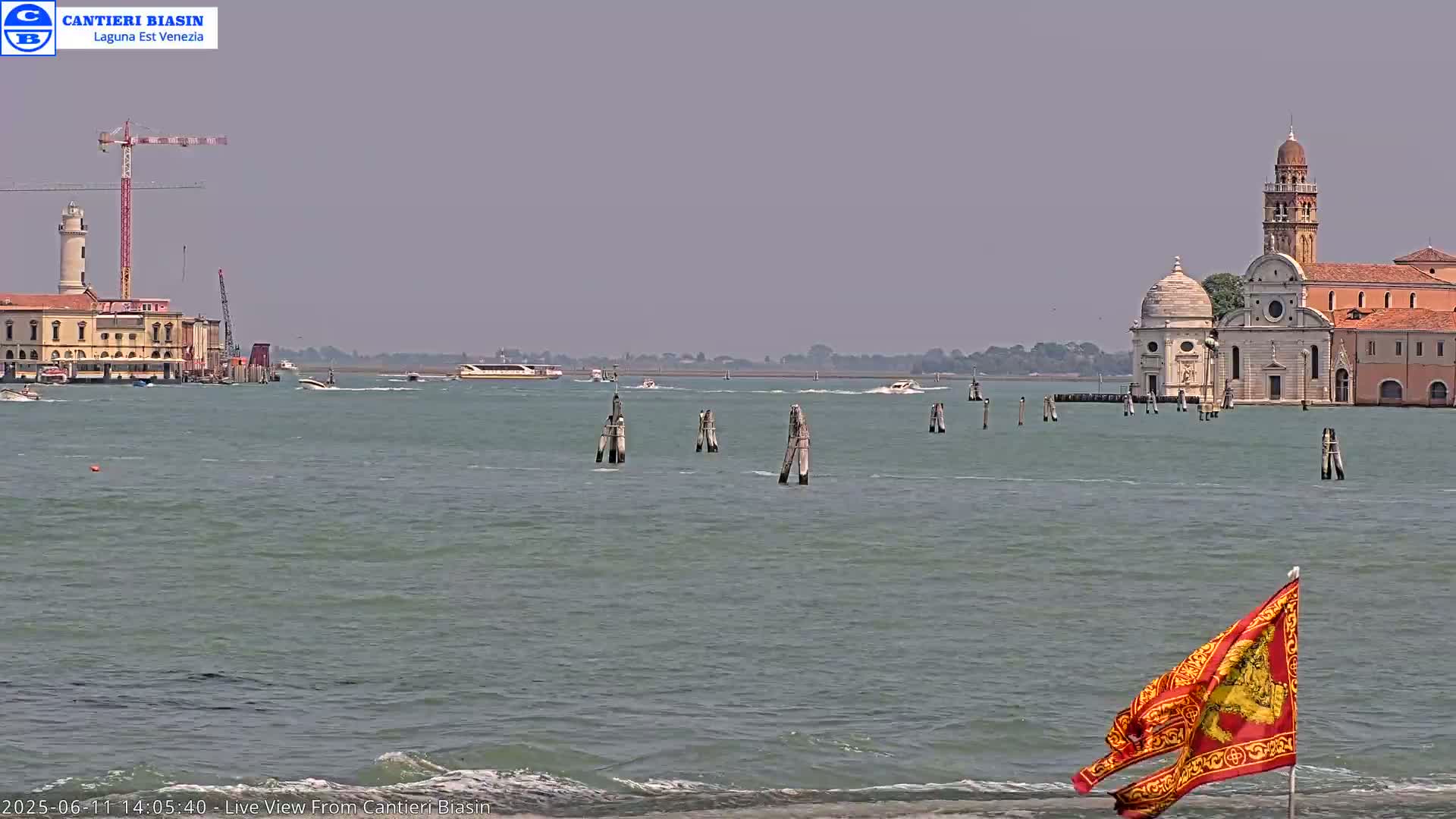 A hazy, sunny day shows a waterway with several boats, a building with a lighthouse and construction crane, wooden posts in the water, a church complex, and a flag on a shore in the foreground.