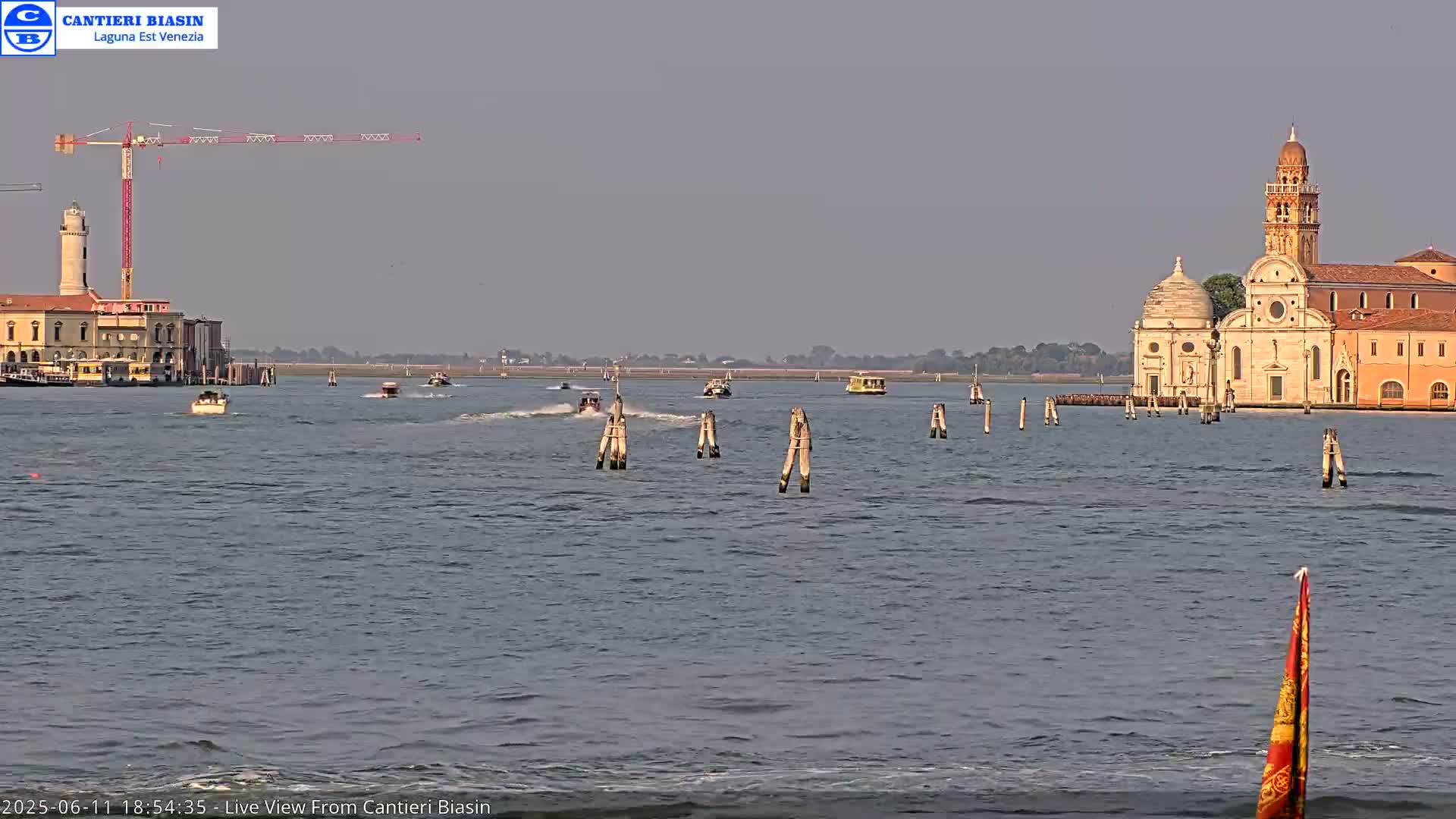 A hazy day shows a calm body of water with several small boats, wooden posts, a lighthouse, a construction crane, and a large church-like building in the distance.