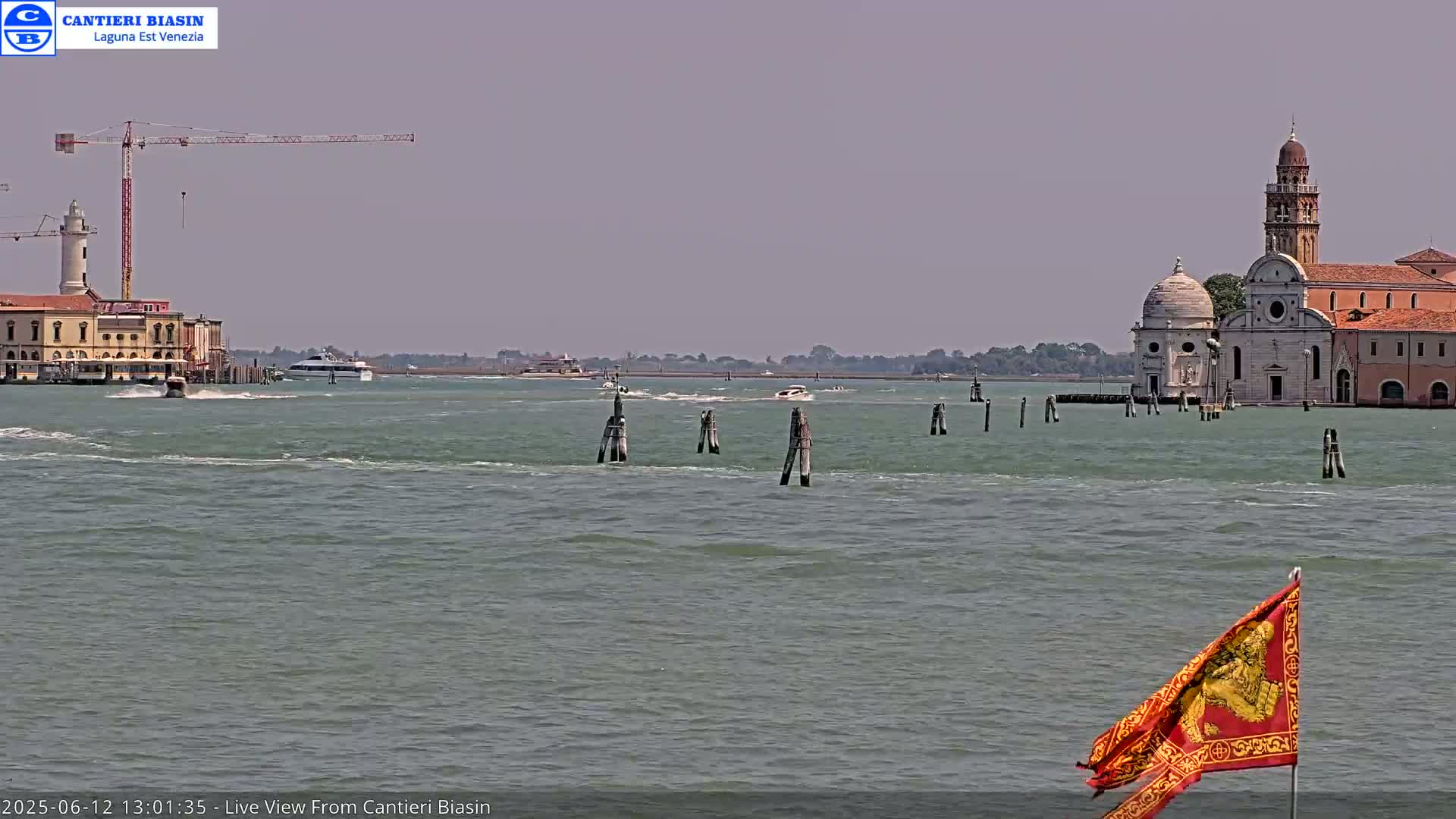 A calm, hazy day shows a Venetian waterway with boats, buildings, a construction crane, and a flag in the foreground.