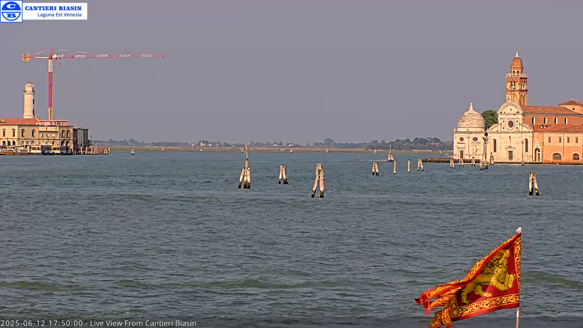 A calm body of water displays a red flag with a gold emblem, a distant church with a bell tower, a building with a lighthouse, and a construction crane under a hazy sky.