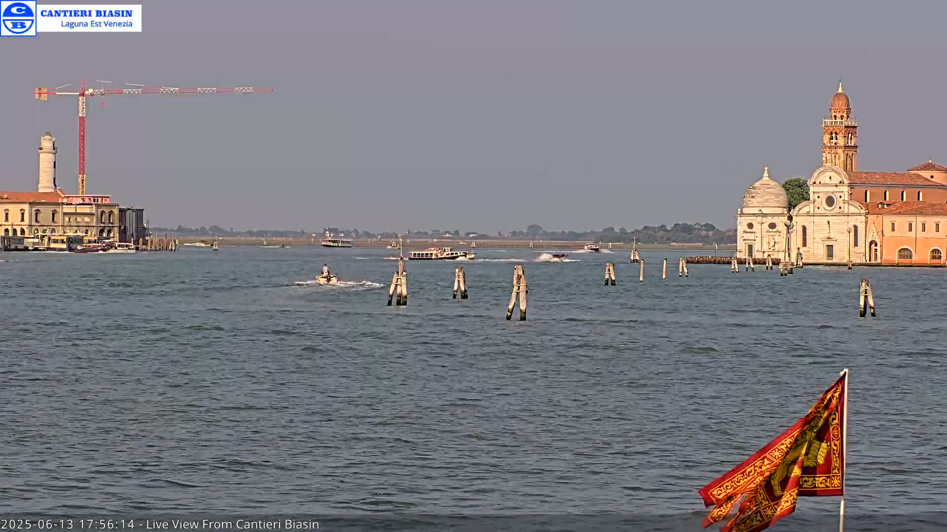 A calm body of water shows several boats, a construction crane, a lighthouse, and a church on the horizon under a hazy sky.