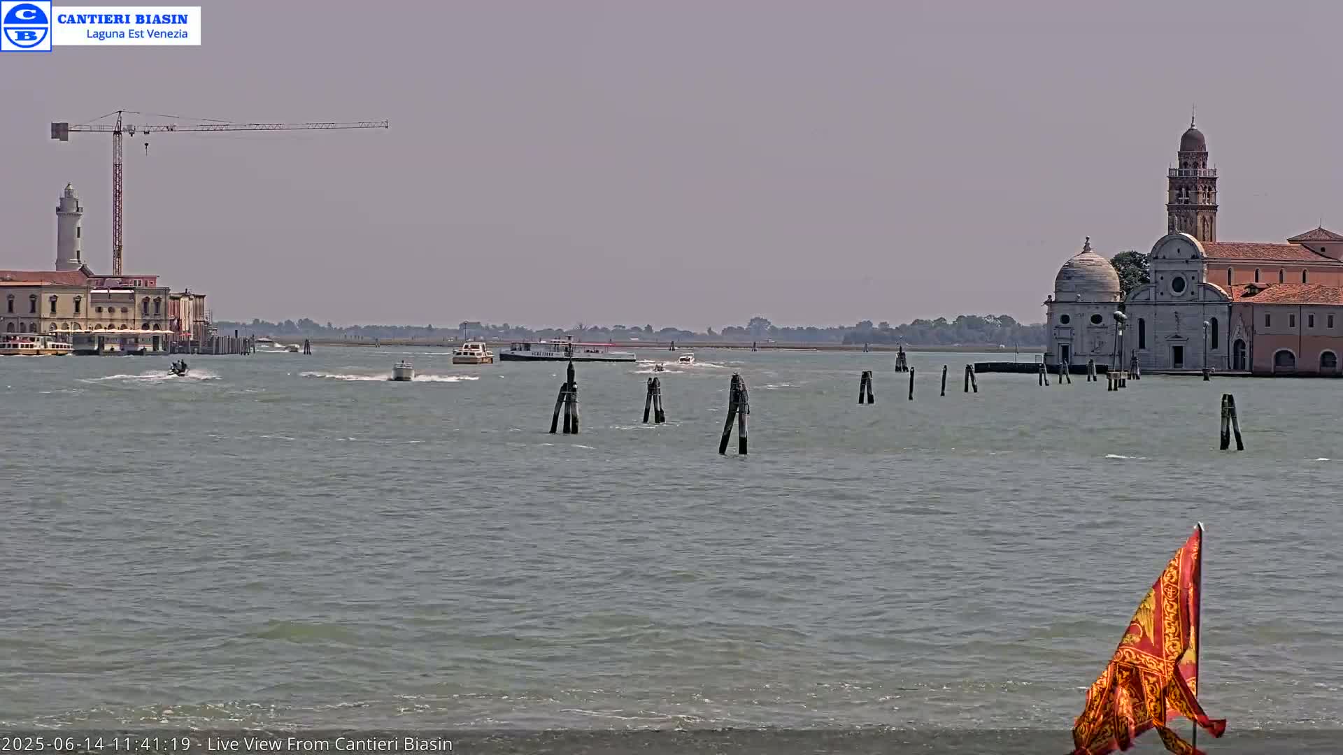 A hazy day shows a calm body of water with several boats, a crane, a lighthouse, and a church complex in the distance.