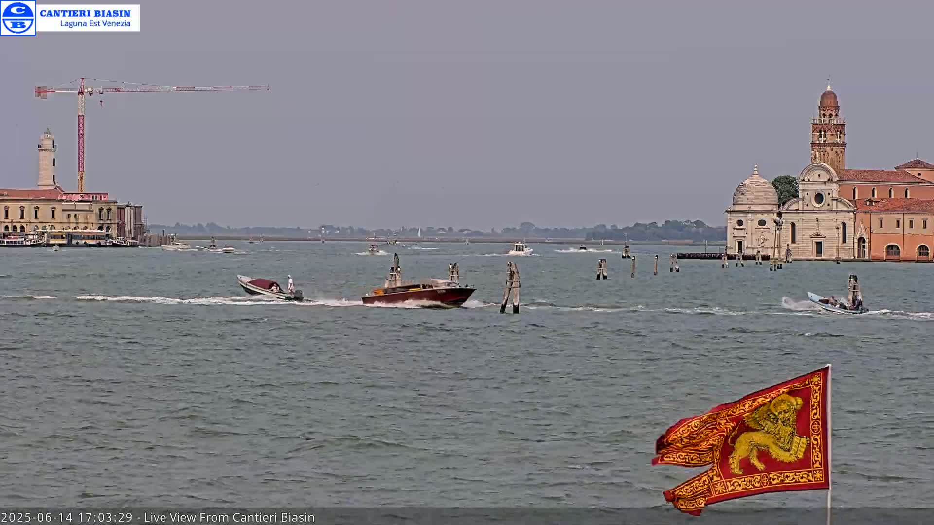 Several motorboats travel across a body of water toward a church and other buildings on a landmass in the distance, under an overcast sky.