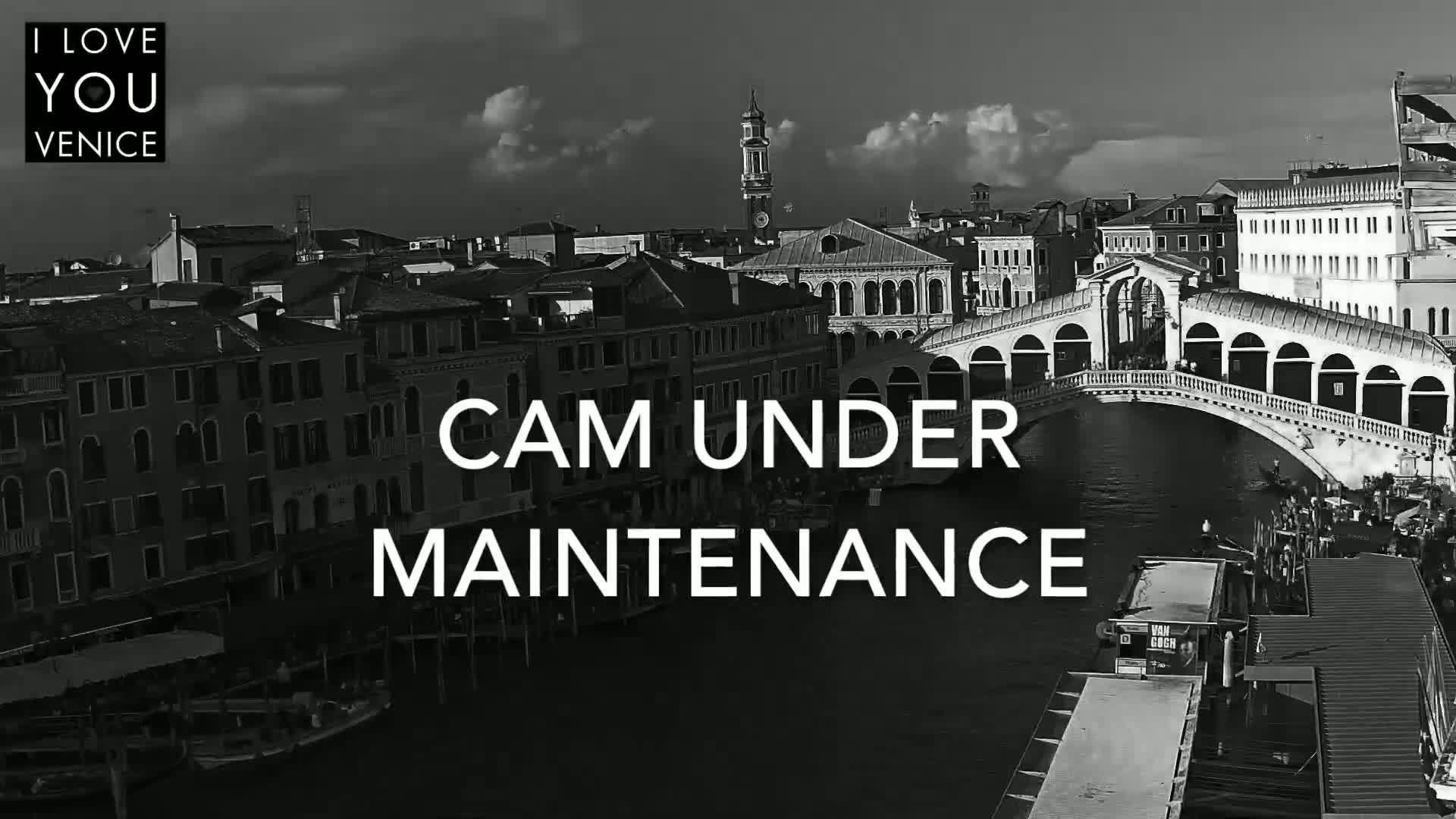 A black and white, high-angle view shows a canal in Venice, with a bridge and numerous buildings under a partly cloudy sky.