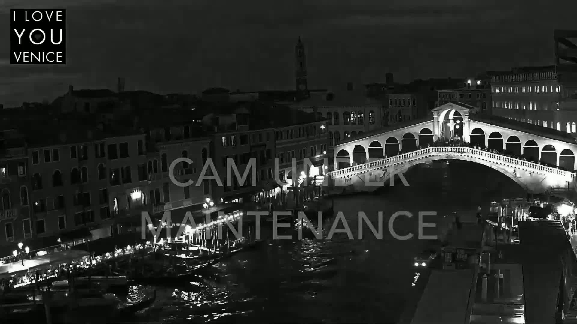 A nighttime black and white view of a bridge over a canal in Venice, with boats and buildings lining the waterway.