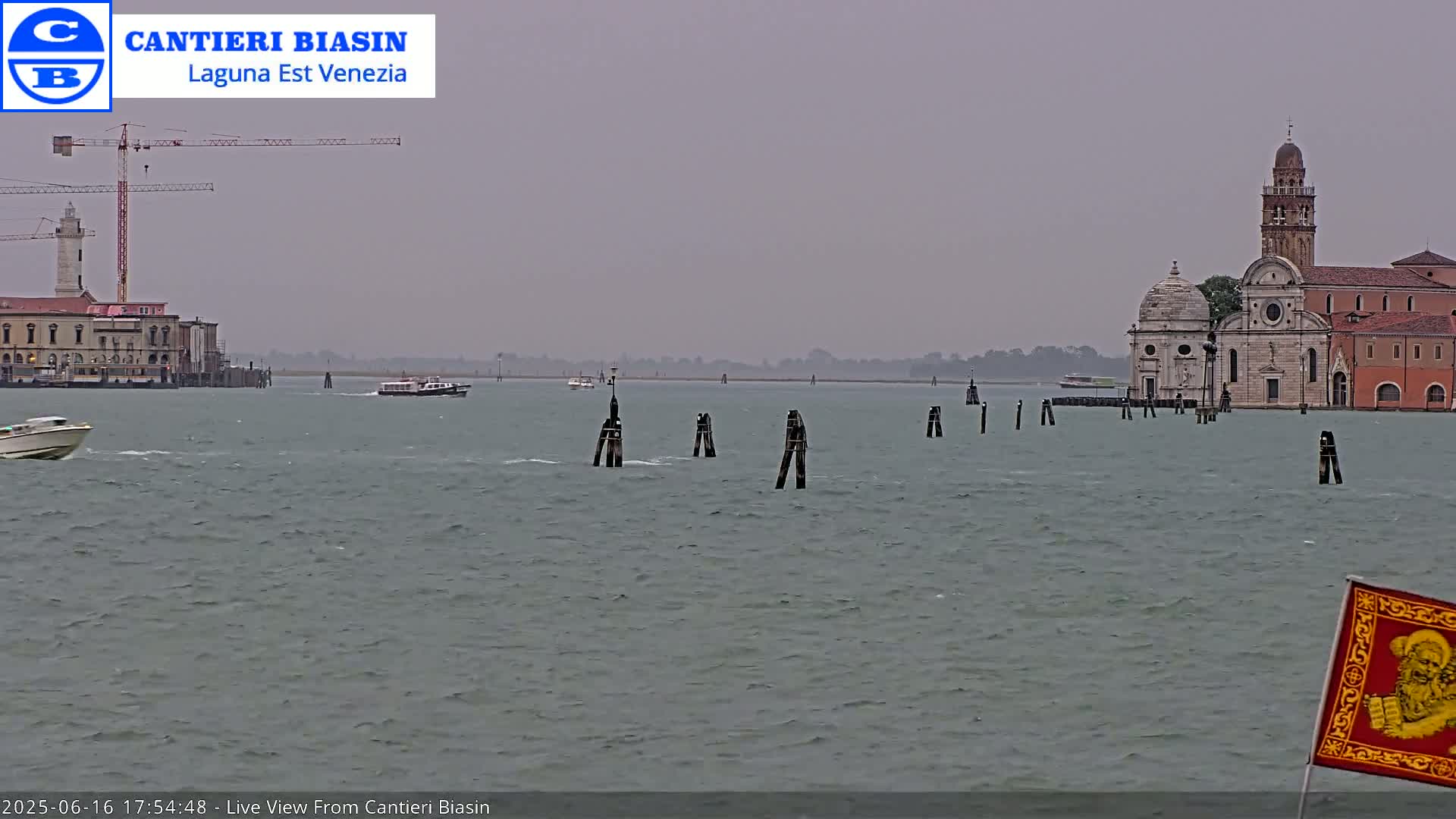 A hazy, overcast day reveals a waterway with boats, a distant church, and a construction crane on the left, all framed by several wooden posts in the water.