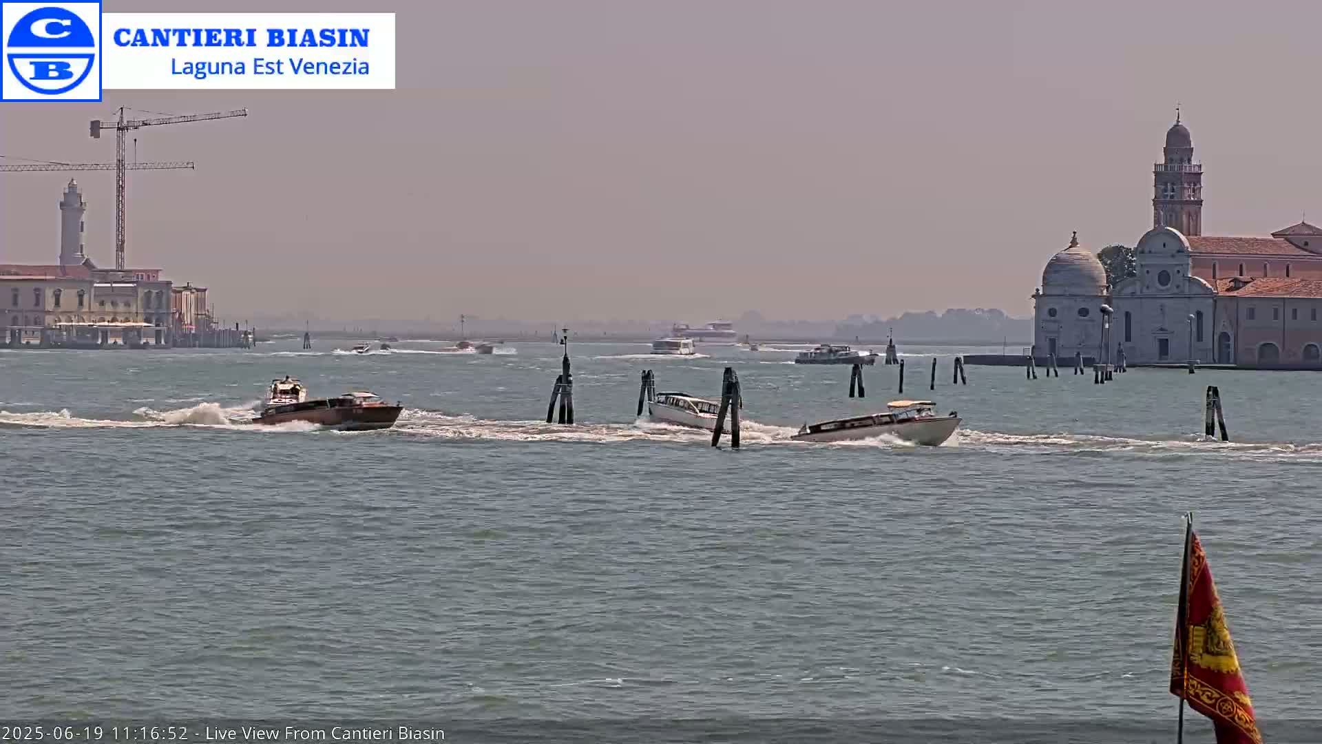 Several motorboats travel across a calm, hazy waterway toward a distant island with buildings, including a church with a bell tower.