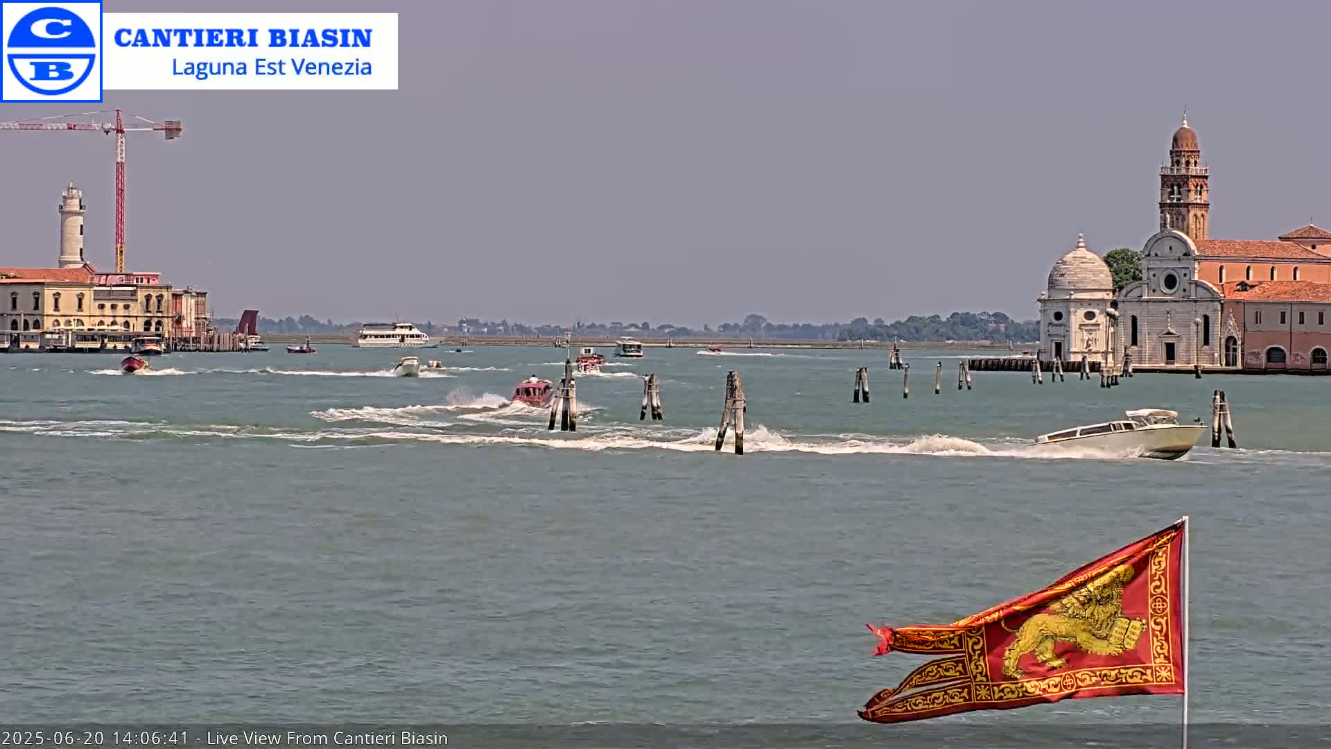 Several boats travel across a body of water on a sunny day, passing near a Venetian-style building and a flag with a lion emblem.