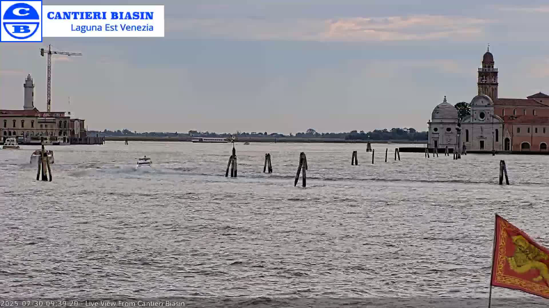 A calm, overcast day shows a waterway with boats, wooden posts, buildings including a church and a lighthouse in the distance.