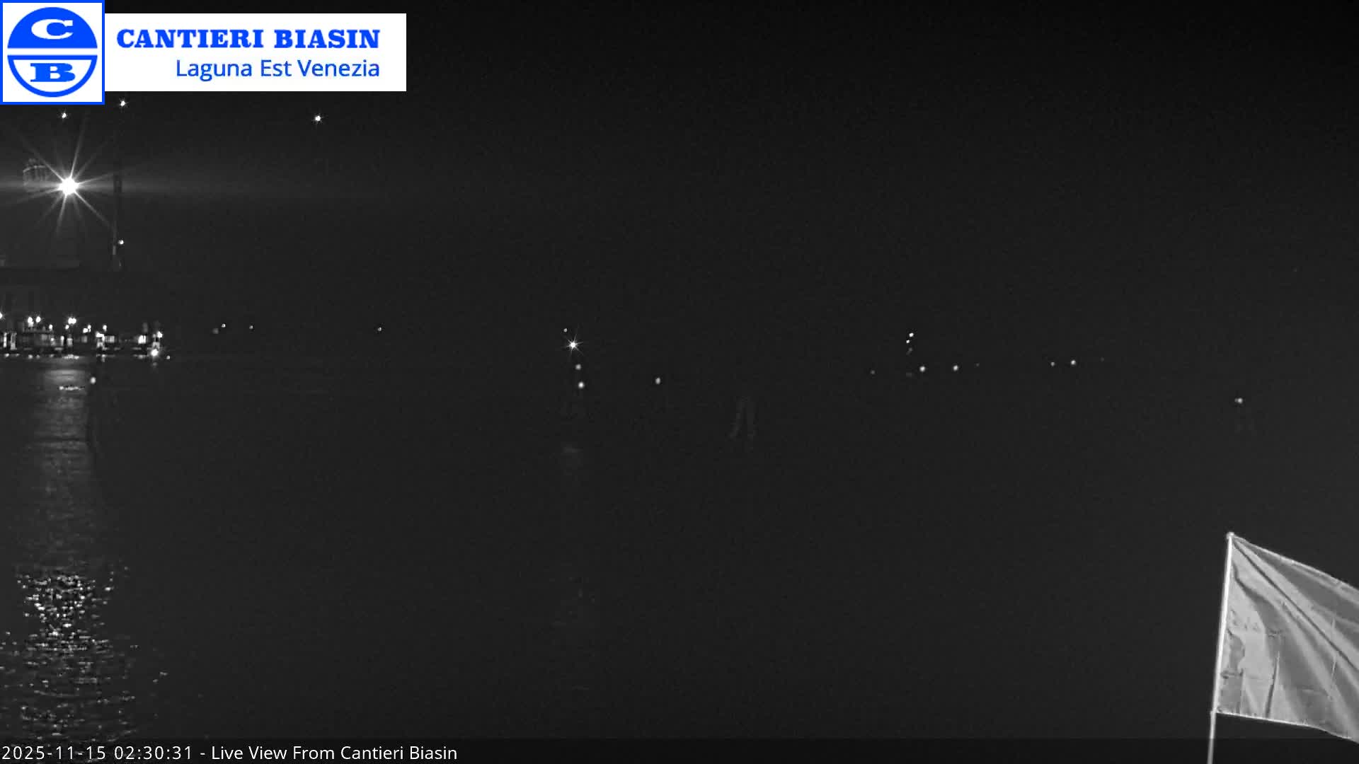 A clear, dark night scene overlooks a body of water, featuring a brightly lit structure reflecting on the left, scattered distant lights along the horizon, and a flag visible in the bottom right corner.