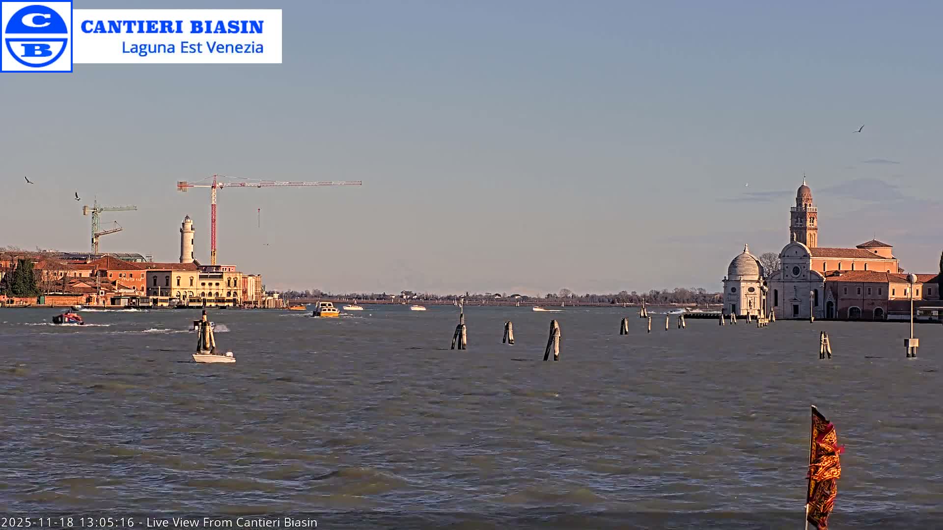 A clear, dark night scene overlooks a body of water, featuring a brightly lit structure reflecting on the left, scattered distant lights along the horizon, and a flag visible in the bottom right corner.