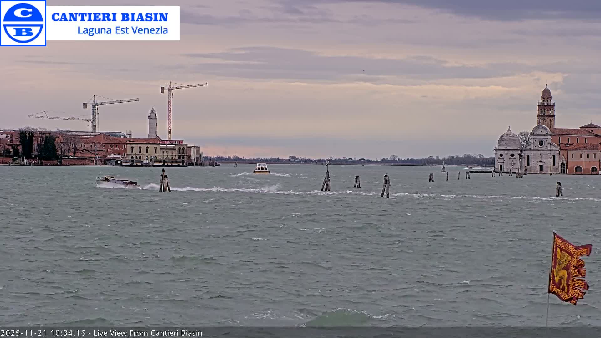 Choppy gray-green water with two boats cutting wakes and numerous navigation posts reflects an overcast sky, flowing between a left bank featuring construction cranes and a lighthouse, and a right bank with historic buildings including a prominent church with a bell tower, with a red and gold flag flying in the foreground.