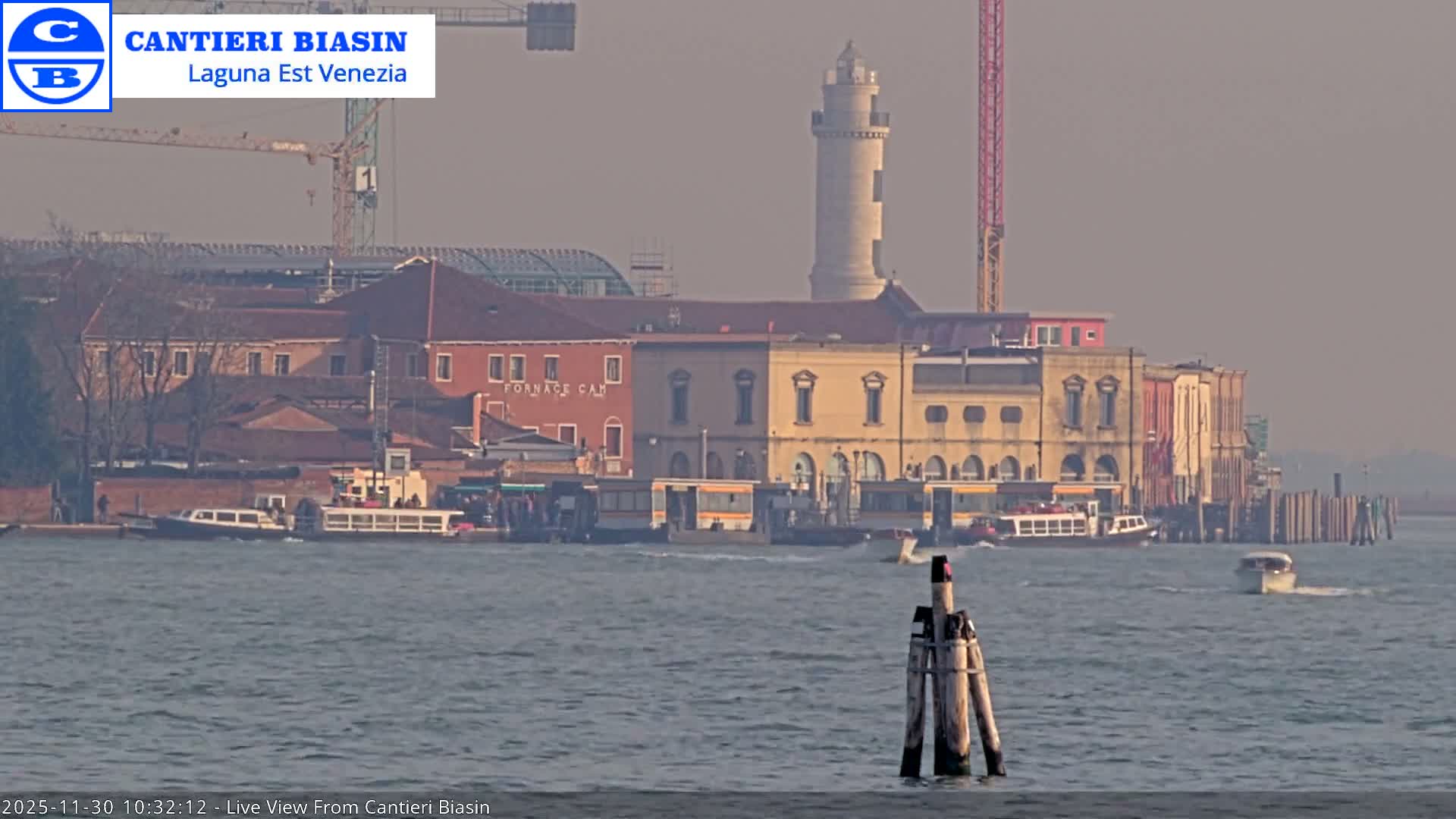 A hazy outdoor scene shows several boats navigating a wide waterway in front of a city shoreline featuring historical buildings, construction cranes, and a prominent lighthouse under a muted sky.