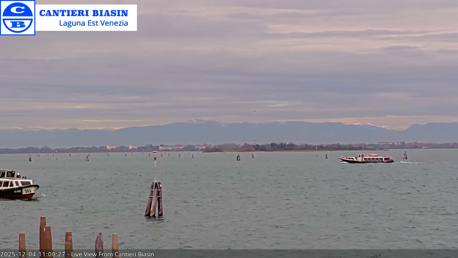 An overcast day reveals a wide lagoon dotted with navigational poles and two boats, a smaller vessel on the left and a larger passenger ferry on the right, set against a distant low-lying shoreline and snow-capped mountains.