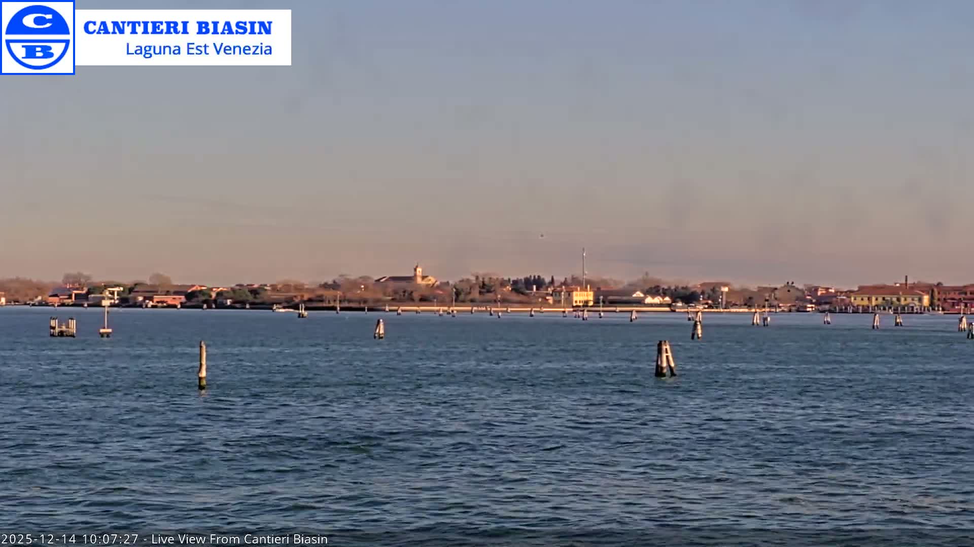 An overcast day reveals a wide lagoon dotted with navigational poles and two boats, a smaller vessel on the left and a larger passenger ferry on the right, set against a distant low-lying shoreline and snow-capped mountains.