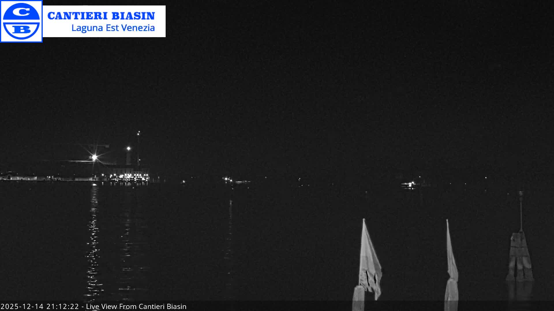 An overcast day reveals a wide lagoon dotted with navigational poles and two boats, a smaller vessel on the left and a larger passenger ferry on the right, set against a distant low-lying shoreline and snow-capped mountains.