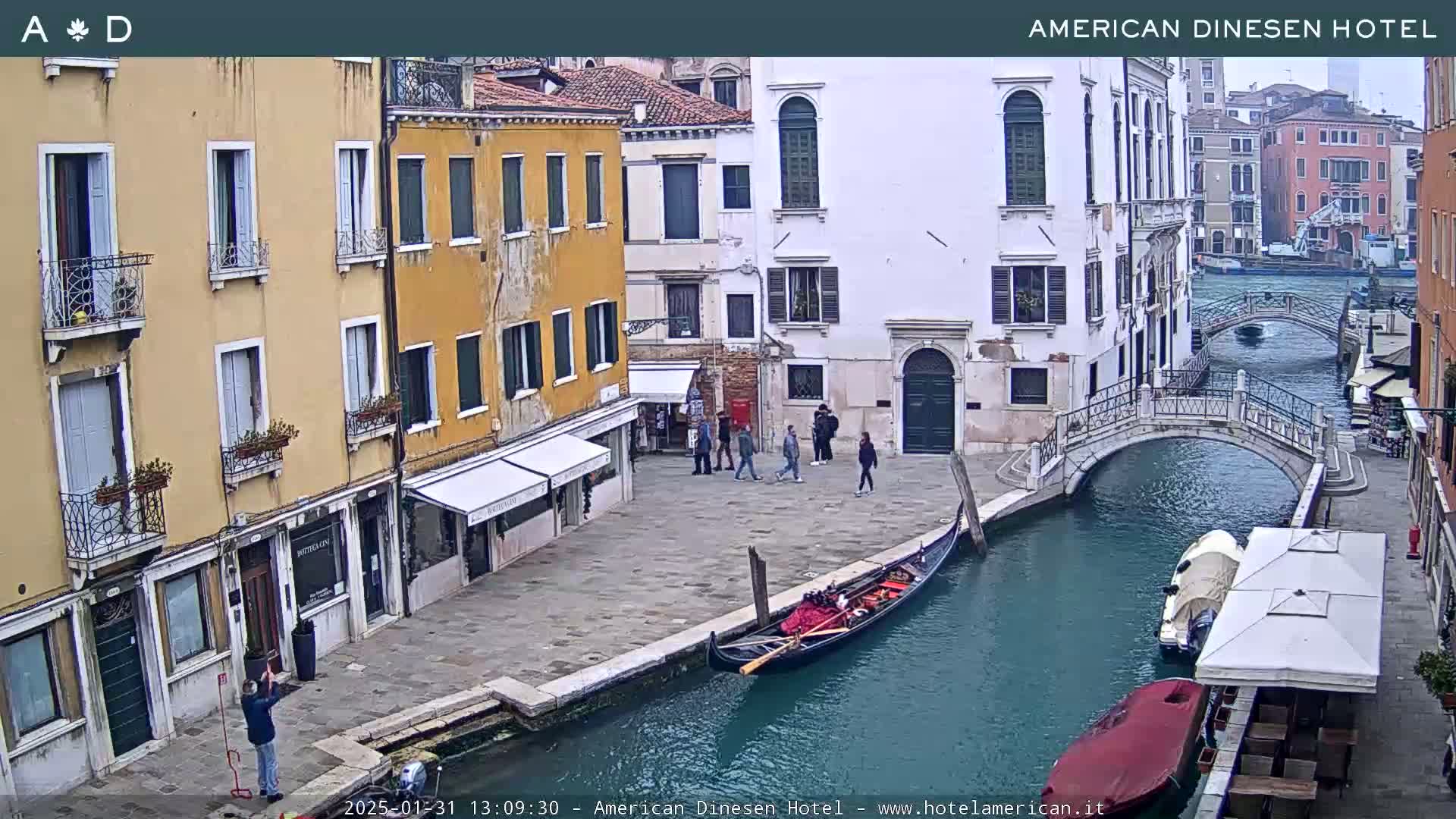 A gondola is moored on a canal in Venice, Italy, under an overcast sky, with pedestrians on a stone walkway and several buildings lining the waterway.