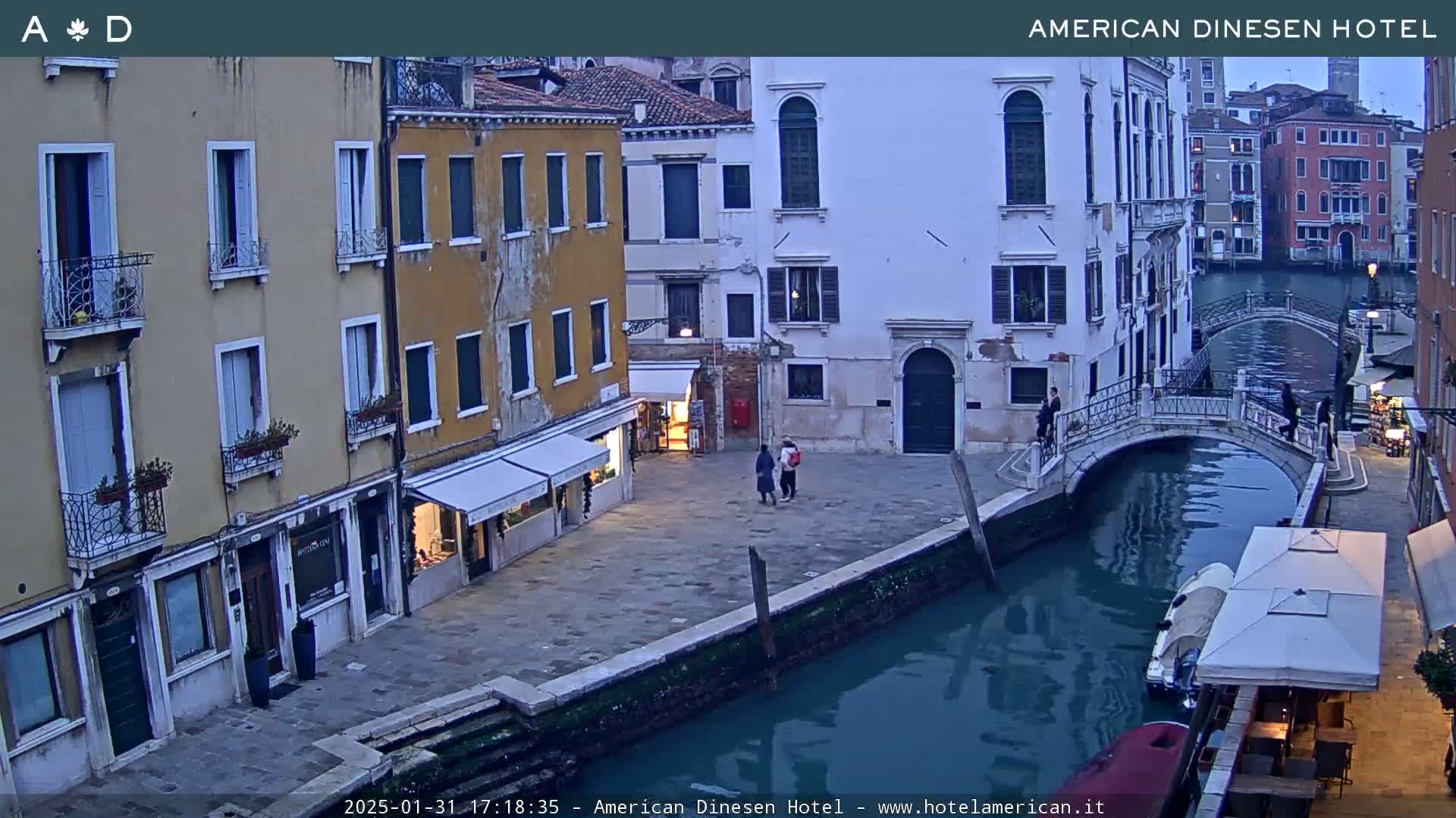 A twilight view of a canal in Venice, showing several buildings lining the waterway, a small bridge, and a few pedestrians.