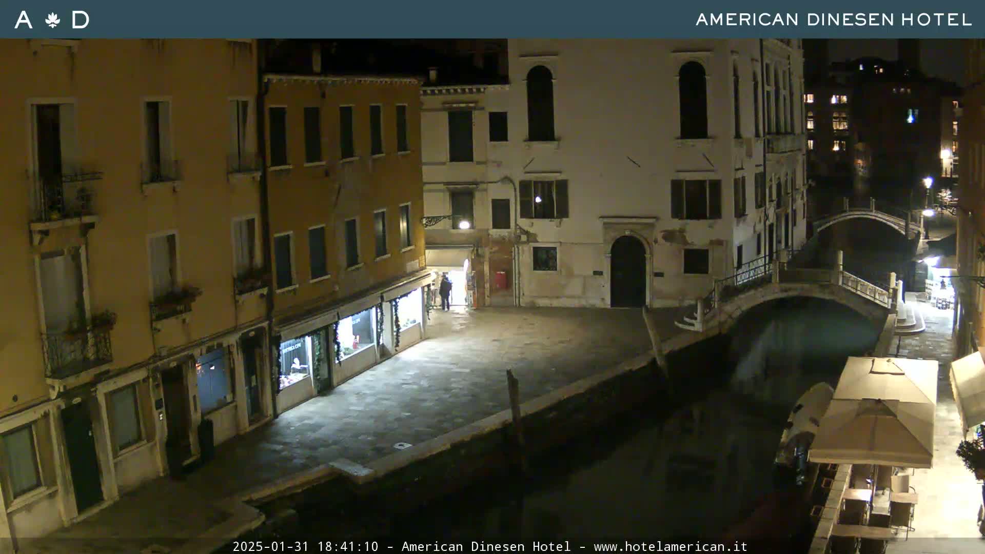 A nighttime view of a canal in Venice, showing several buildings along the waterway and a small bridge, with a patio umbrella visible near the water's edge.