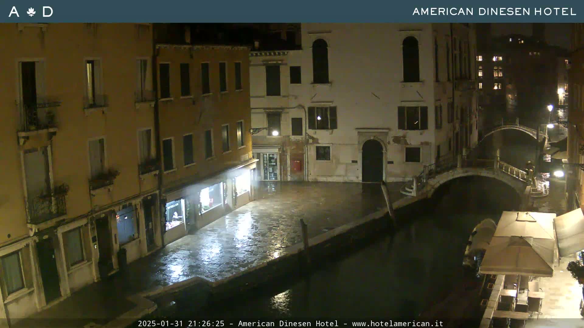 A nighttime view of a canal in Venice, Italy, shows several buildings lining the waterway, with wet pavement reflecting lights and a small outdoor seating area under an awning visible along the canal.