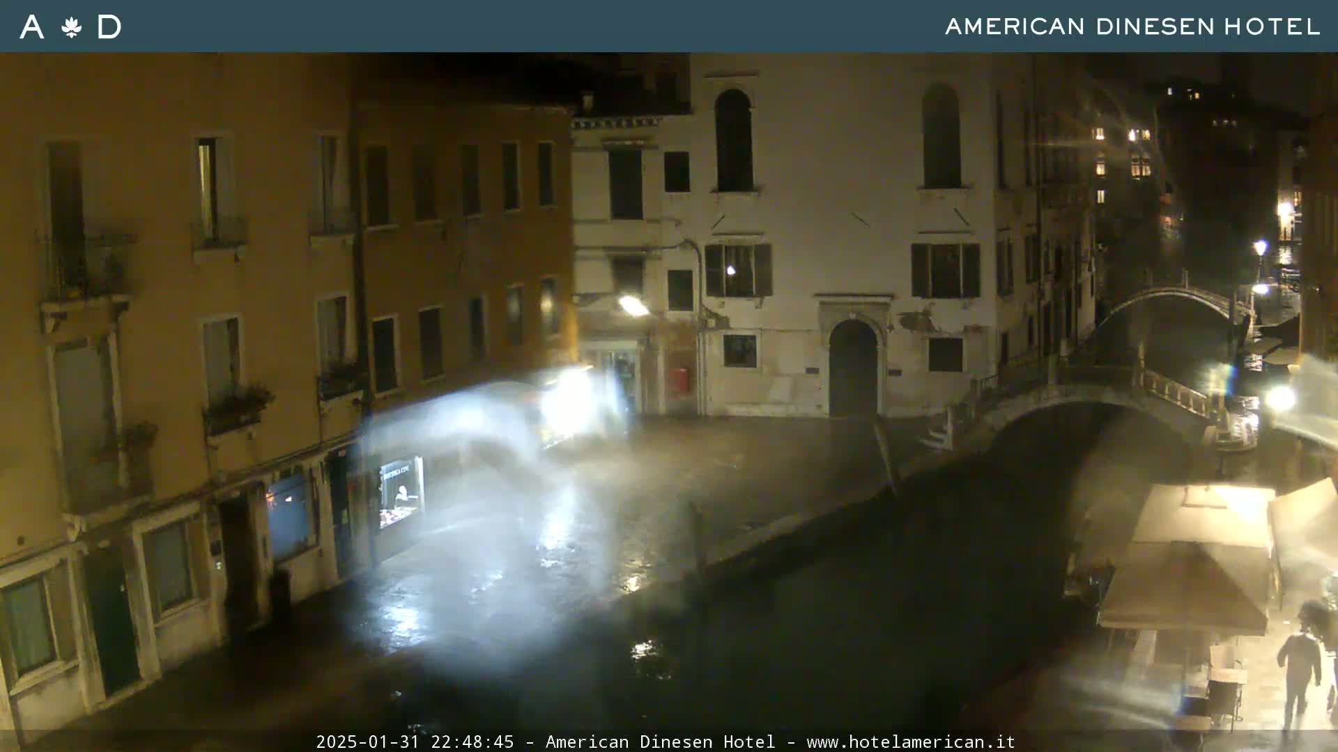 A nighttime view of a canal in Venice, Italy, shows several buildings along the waterway under rainy conditions with water splashing on the ground near the camera.