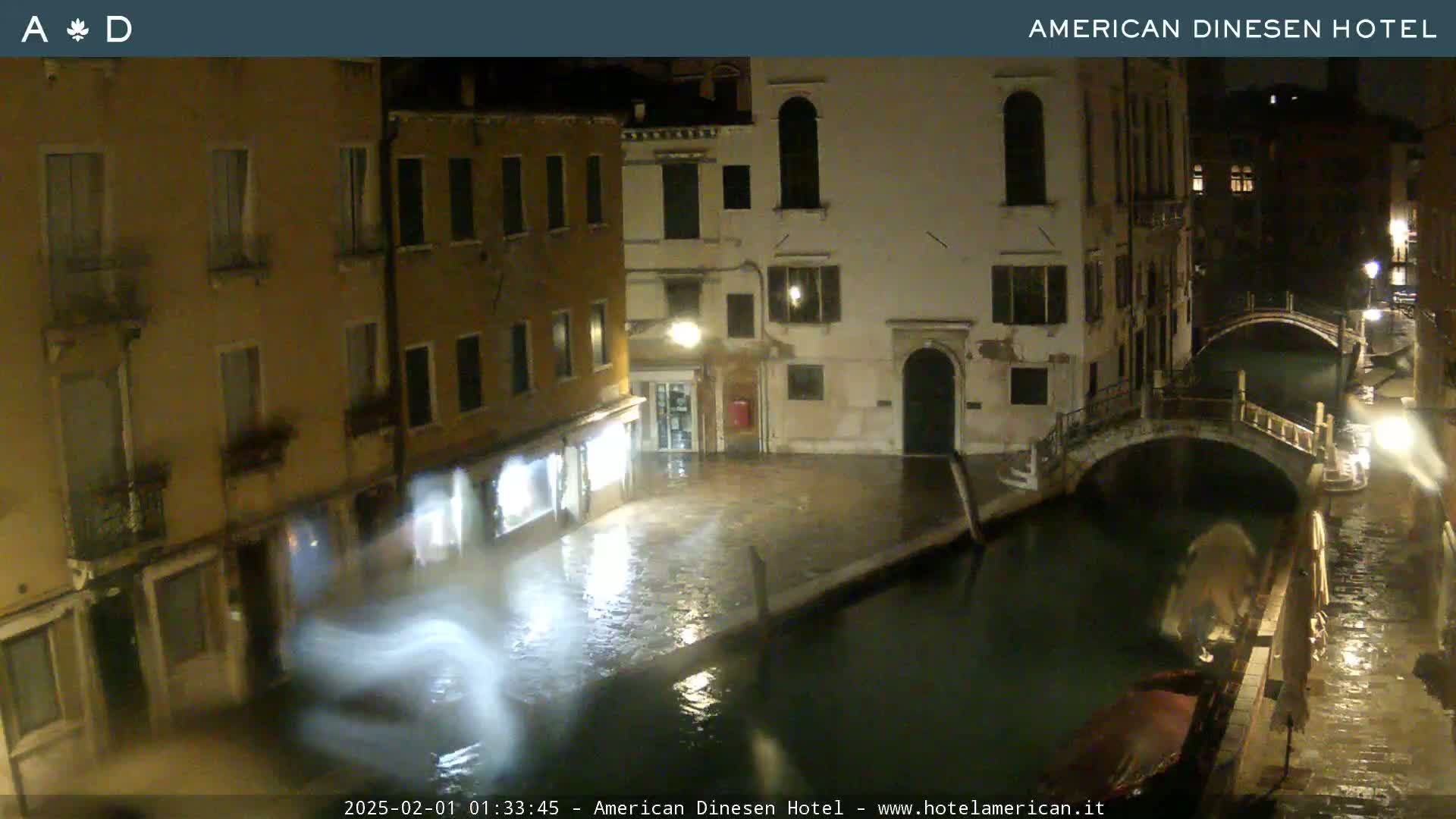 A nighttime view of a canal in Venice, showing buildings lining the waterway and a small bridge, with rain-slicked streets and reflections in the water.